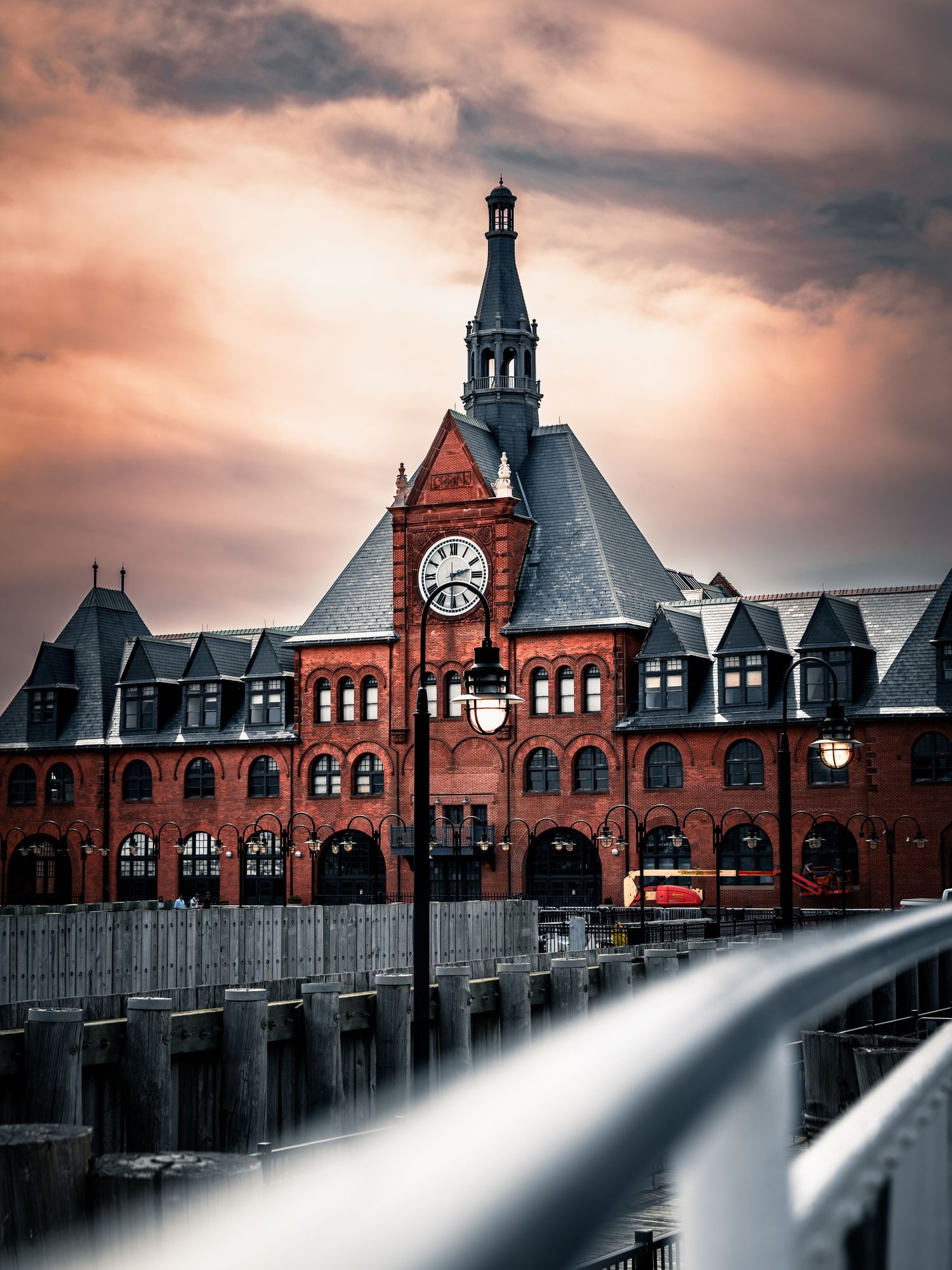 Historic red brick railway station building with clock tower and dramatic sunbeams cutting through moody clouds at sunset.