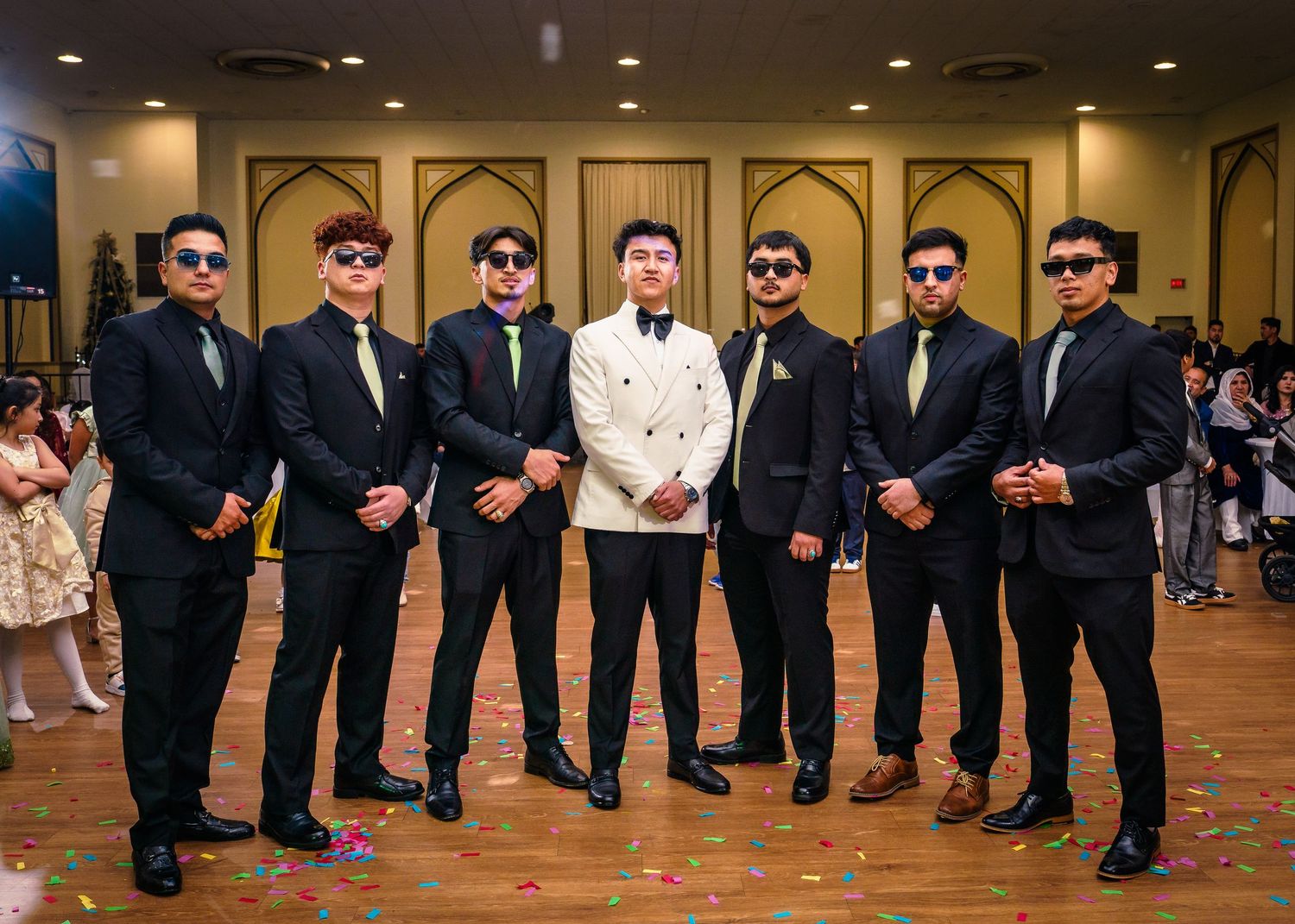 Groomsmen in black suits and sunglasses strike fun poses while lined up in a wedding reception venue.