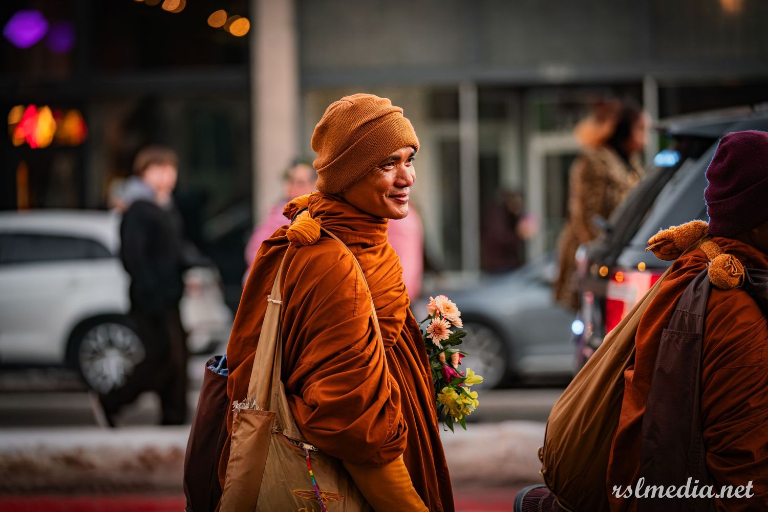 A person in bright orange robes walks on a city street at dusk carrying a brown bag.