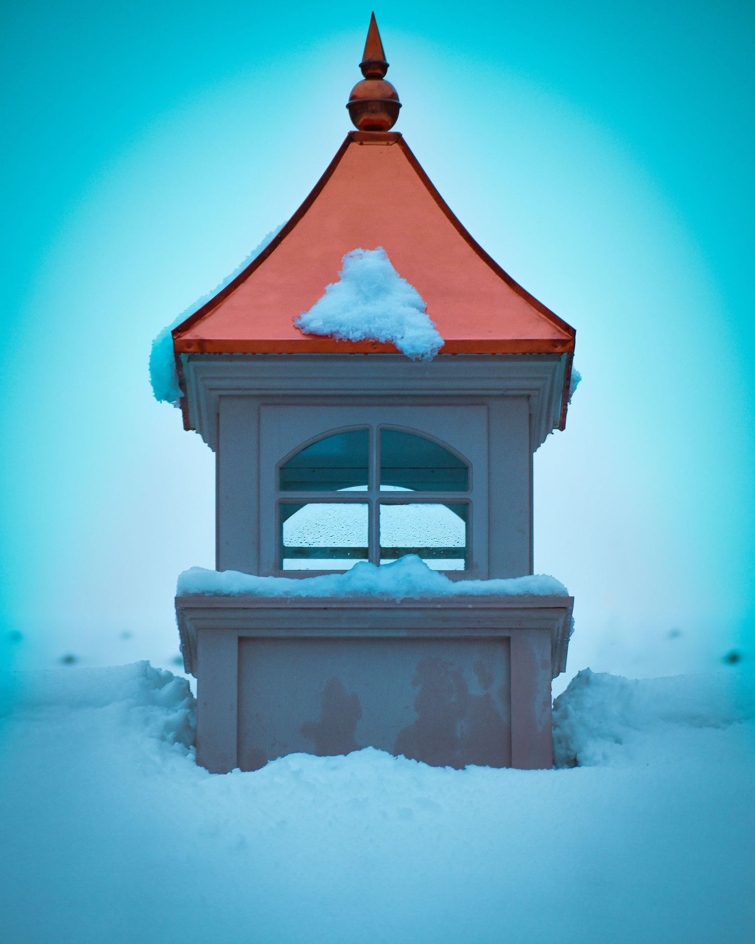 A red-roofed structure stands partially buried in deep snow against a bright turquoise sky.