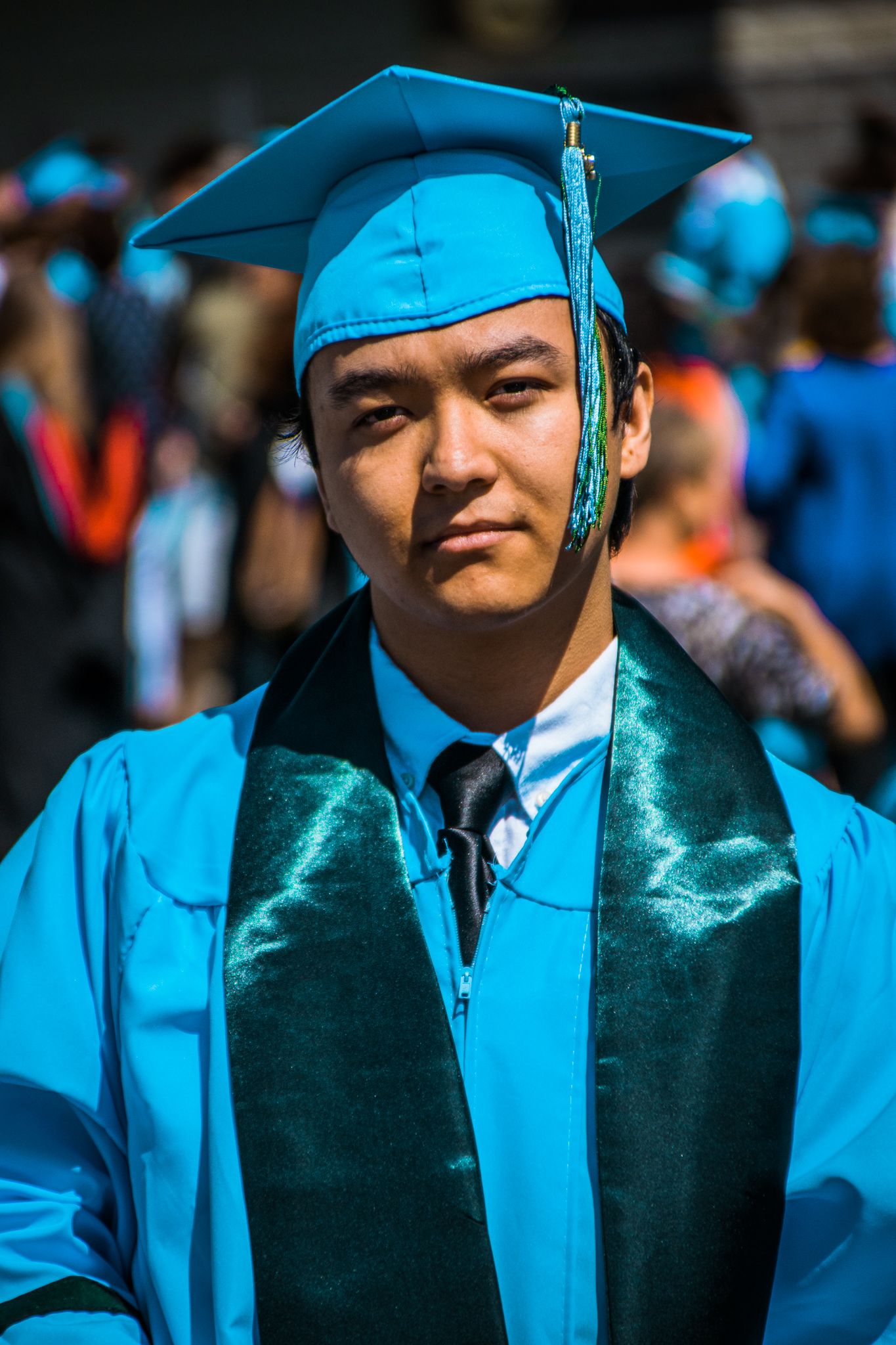 Graduate wearing bright blue cap and gown with green sash during outdoor ceremony.