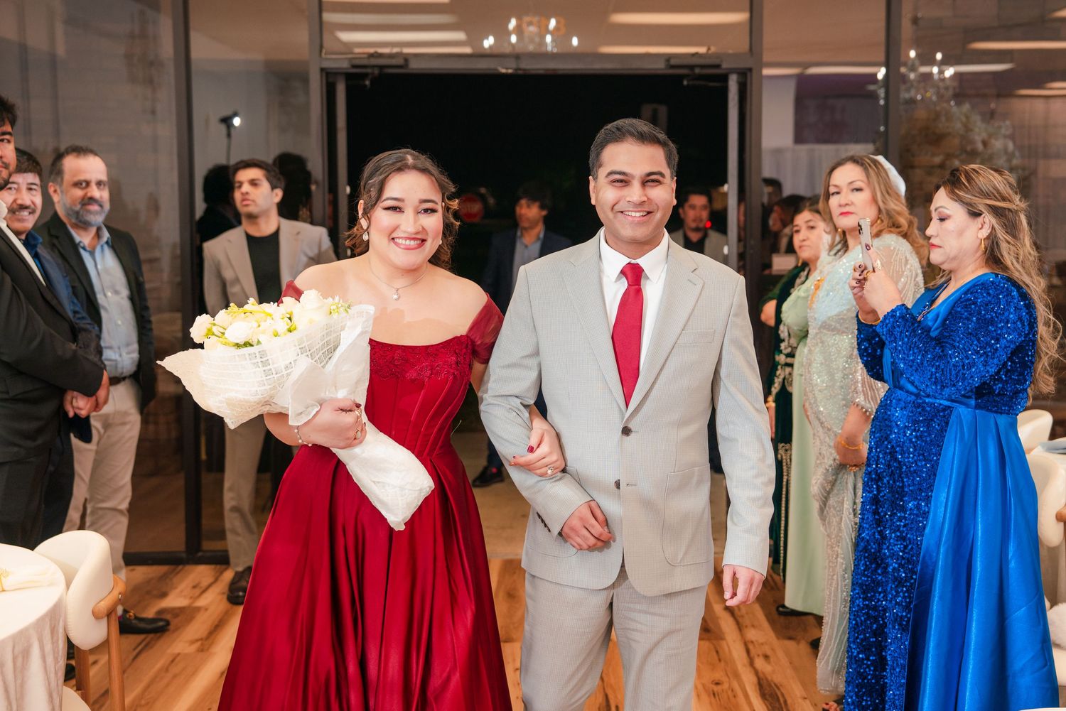 Wedding party members in red and royal blue dresses dance and celebrate at an indoor reception venue.