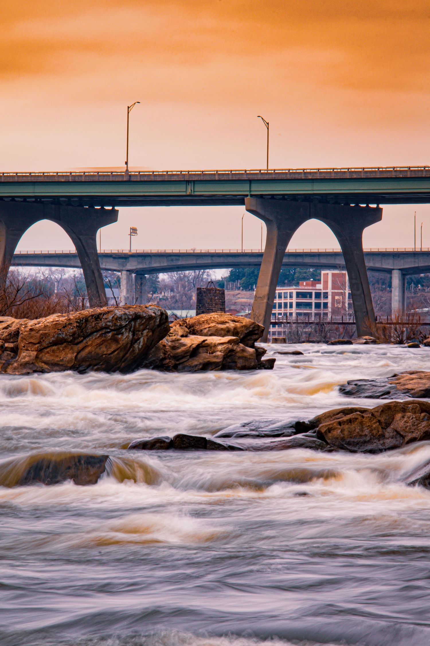 Rushing rapids flow under a bridge during golden sunset with dramatic orange sky.
