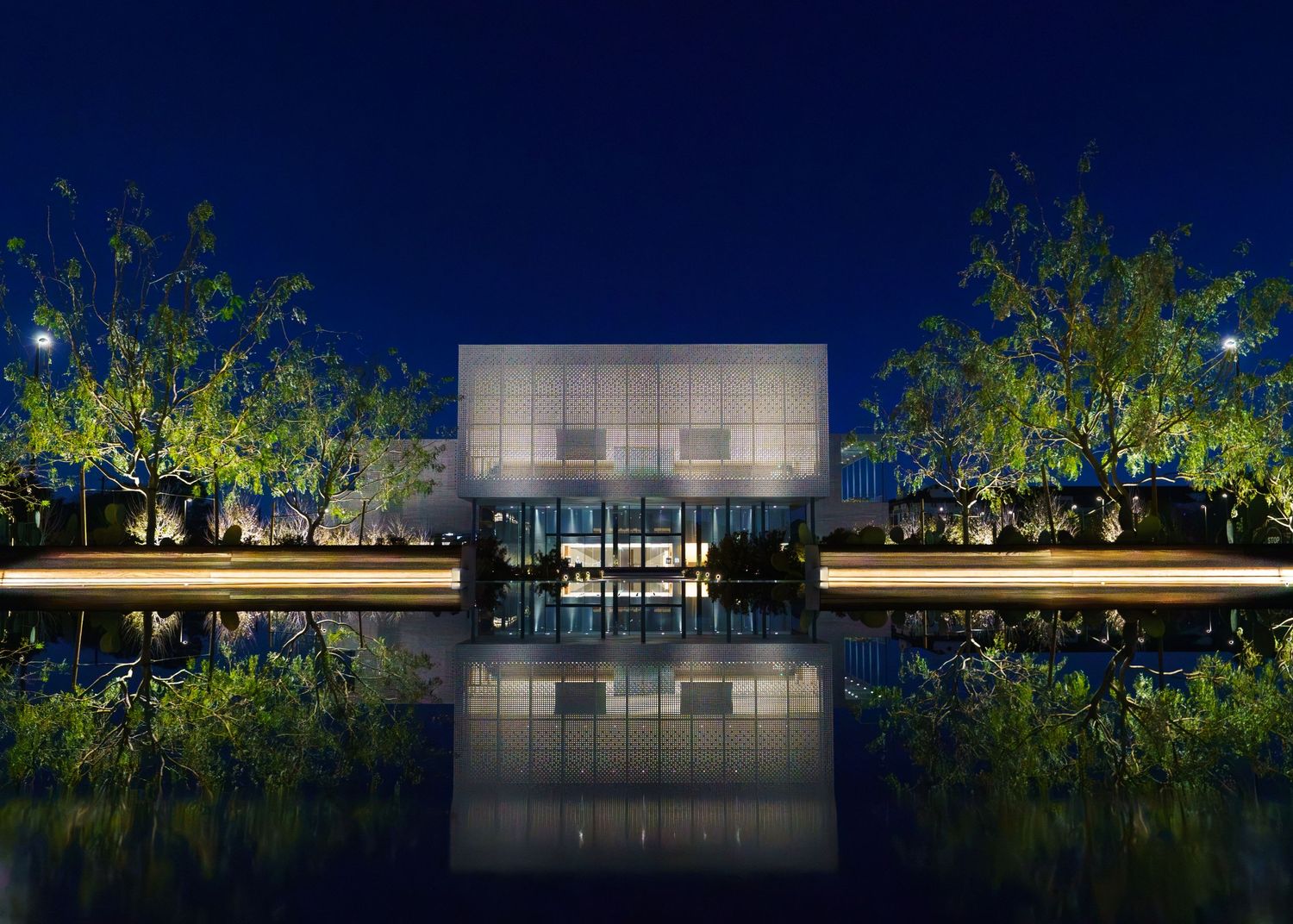 Modern glass building illuminated at night with reflective pond and trees creating a serene architectural landscape.