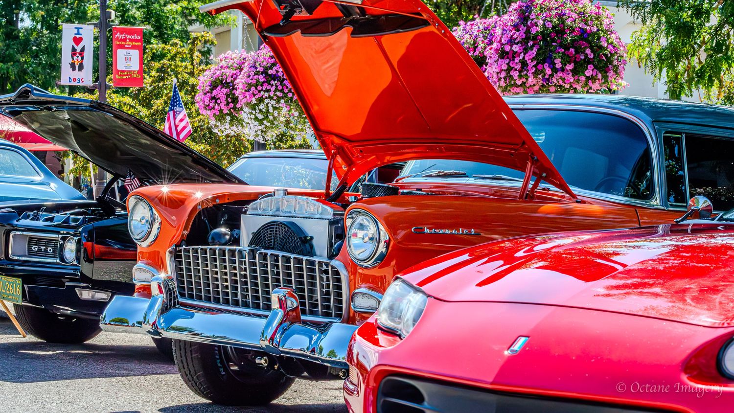 The front end of an orange and silver 1955 Chevrolet Bel Air with a popped hood behind the front end of a red 1998 Camaro.