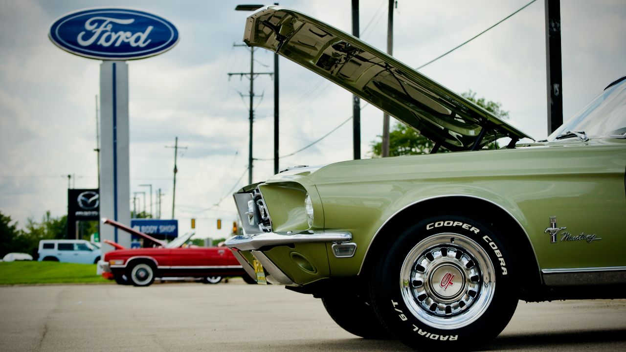 A Seafoam Green 1968 Mustang with an open hood in front of a large Ford logo in the background.