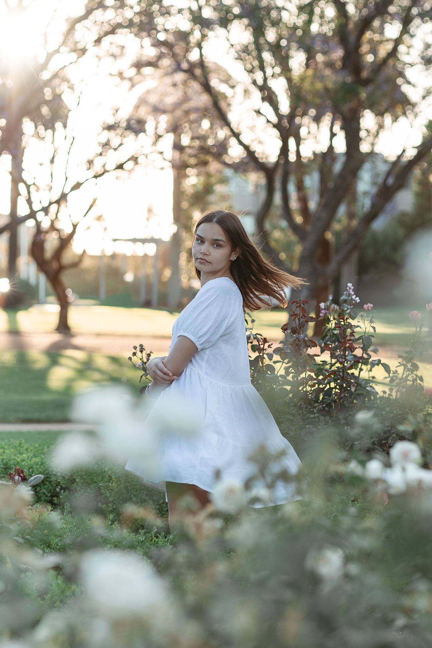 Person in white dress poses among blooming flowers during golden hour.
