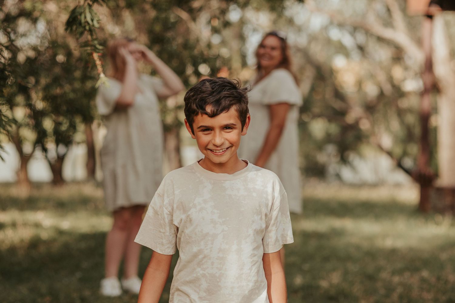 Young person smiling in white t-shirt with blurred figures in background during outdoor gathering.