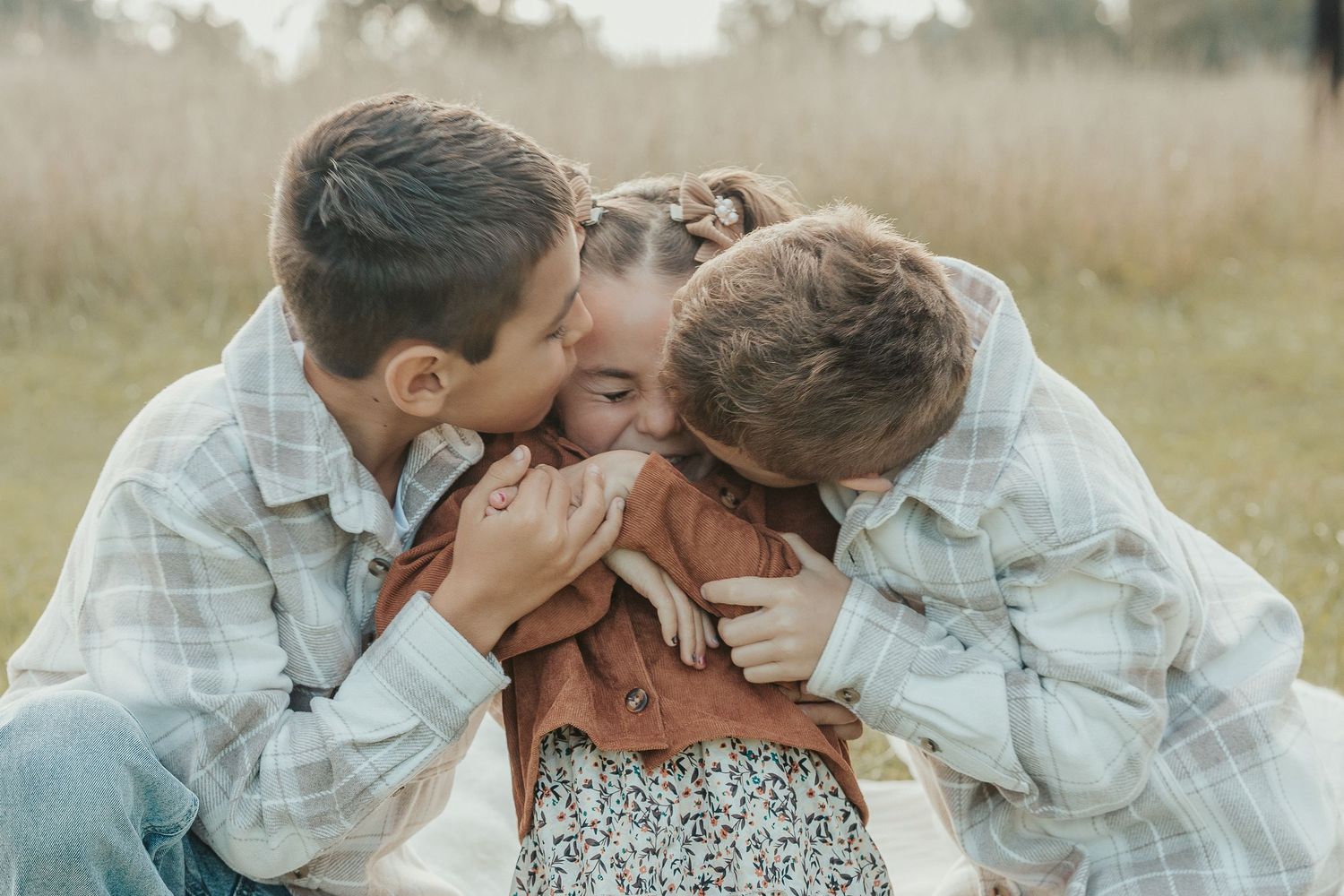 Two children lovingly kiss their mother on the cheeks outdoors in a grassy field during a family photo session.