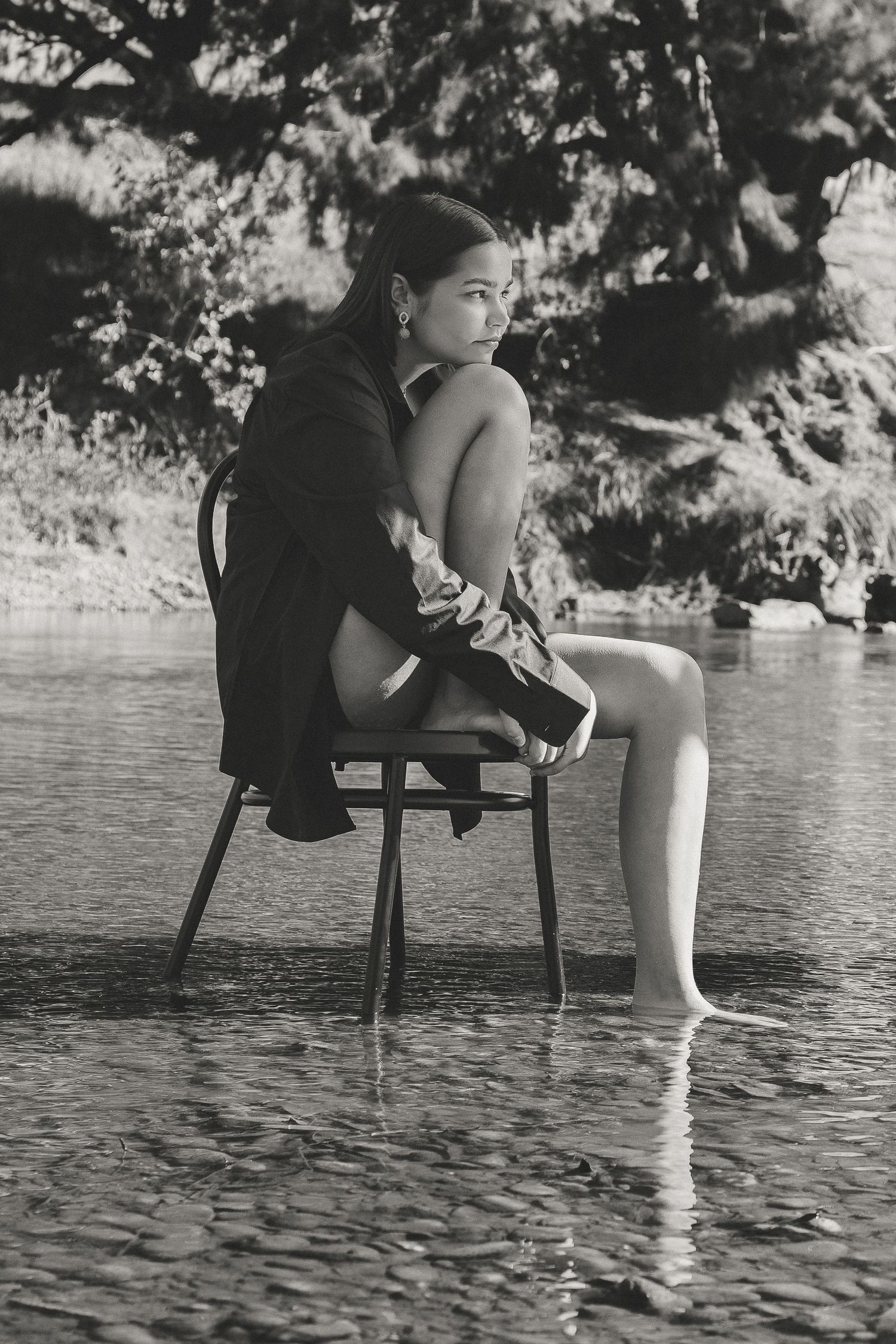 Black and white photo of a young woman sitting on a chair in shallow water, wearing a jacket, with trees in background.