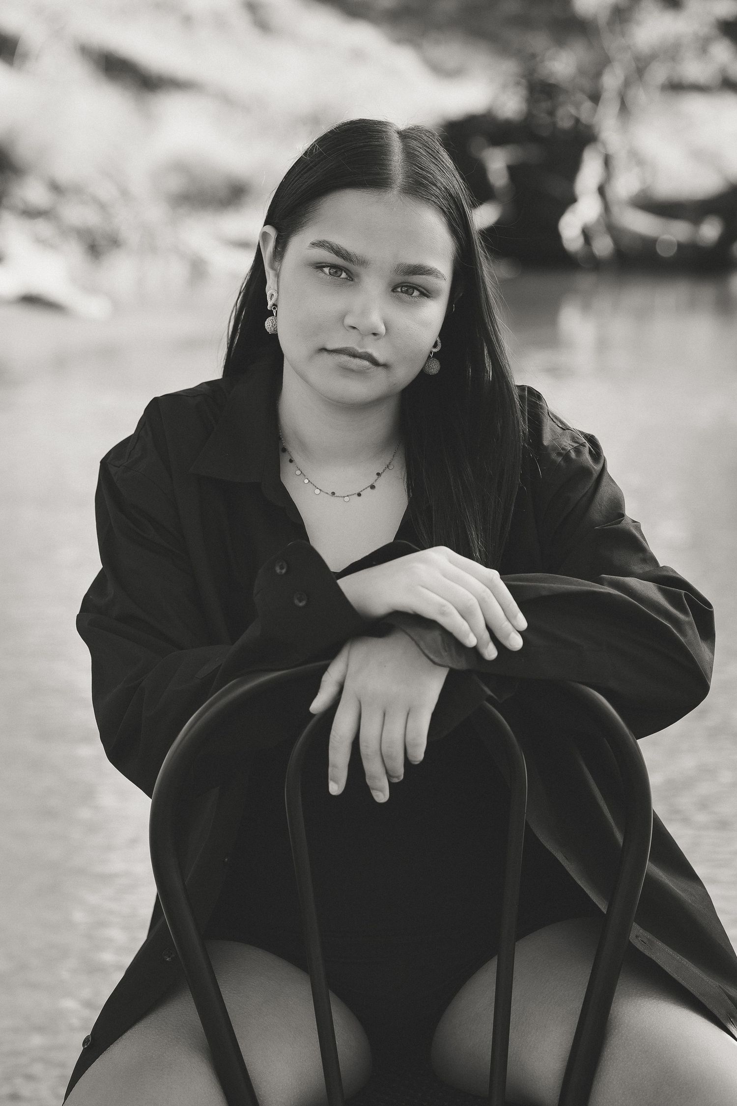Black and white portrait of a young woman in oversized black shirt sitting outdoors near water.