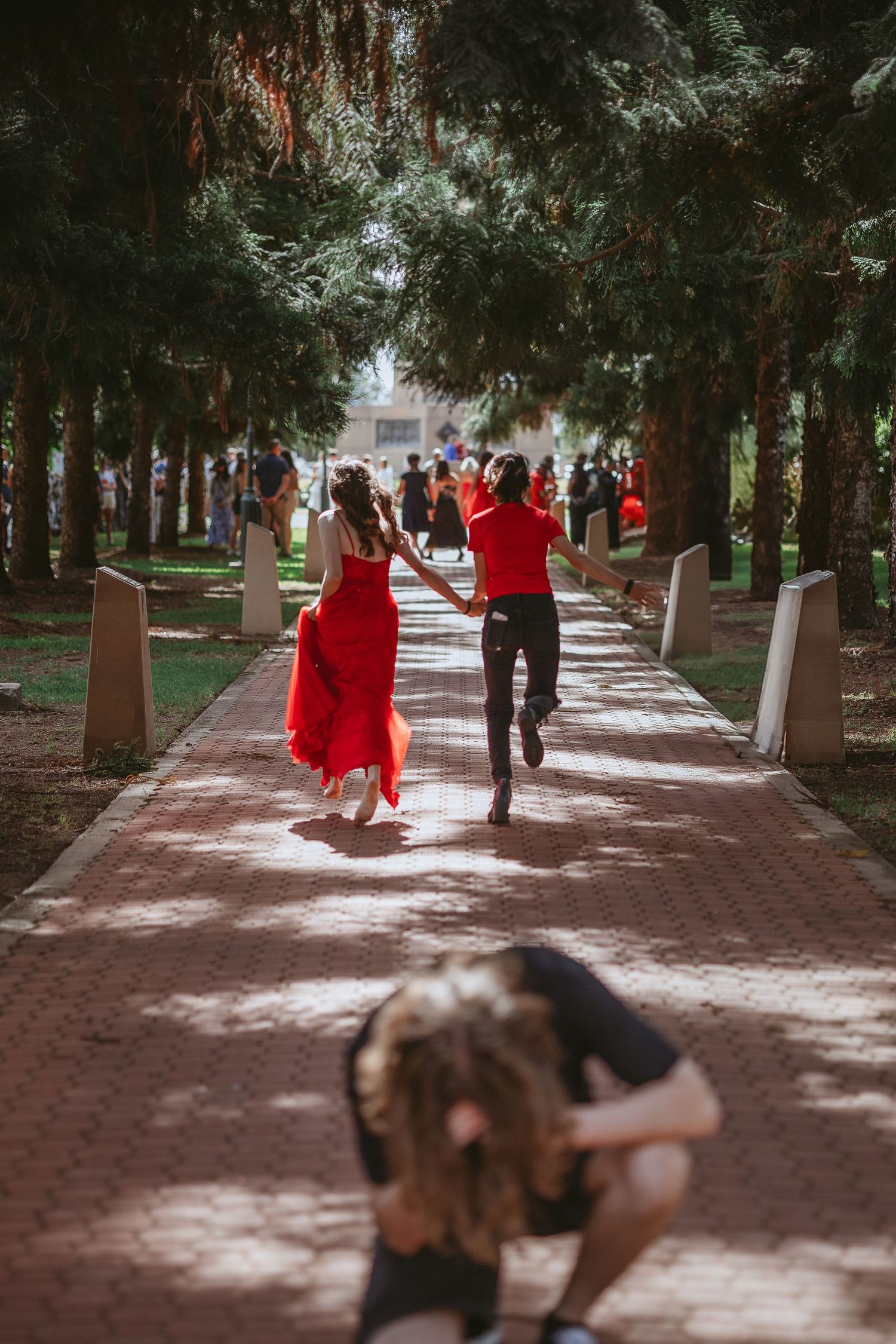 People in red formal wear walking and posing along a tree-lined pathway.