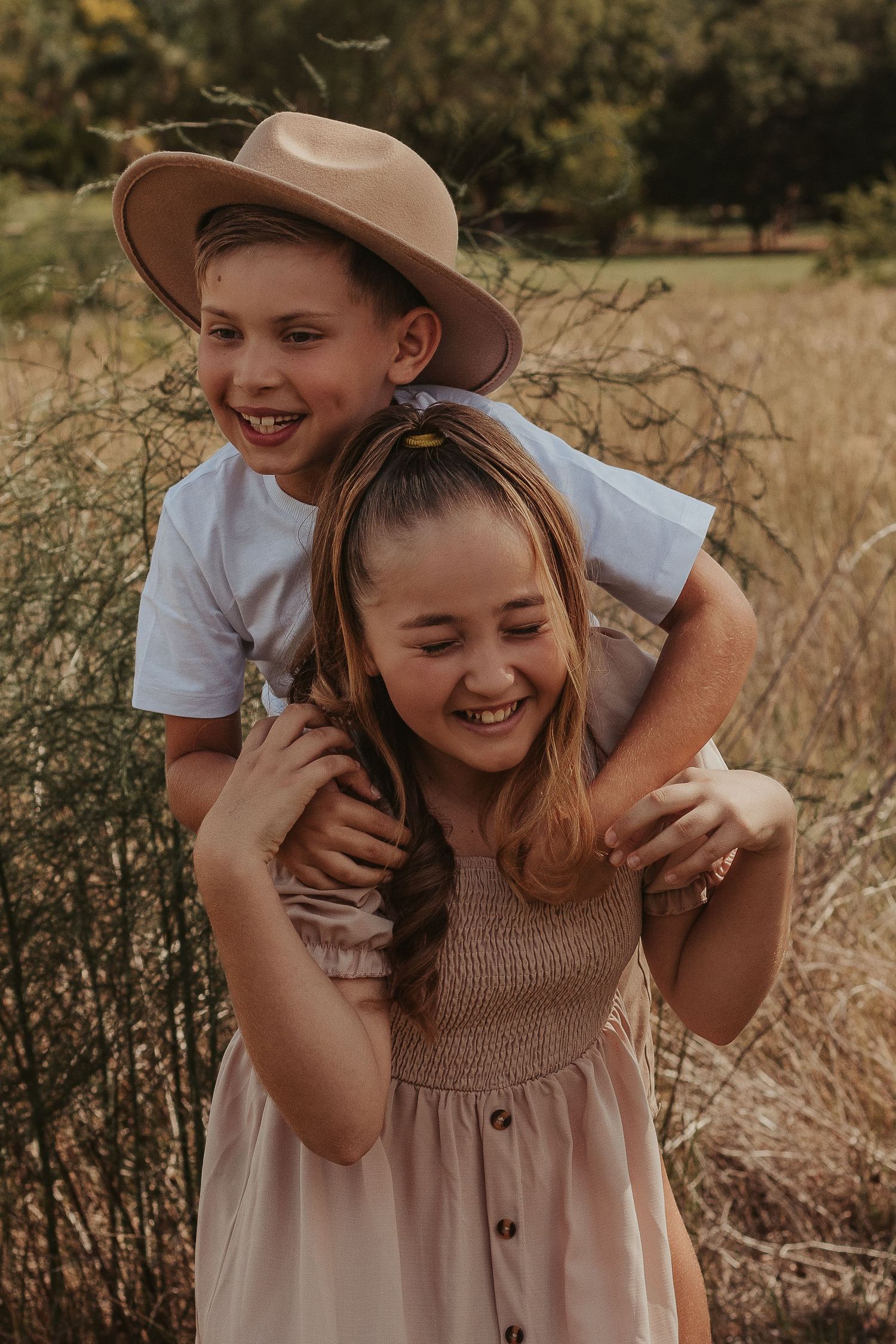 Two smiling children in a sunny field, girl in pink dress giving piggyback ride to boy in white tee and tan hat.