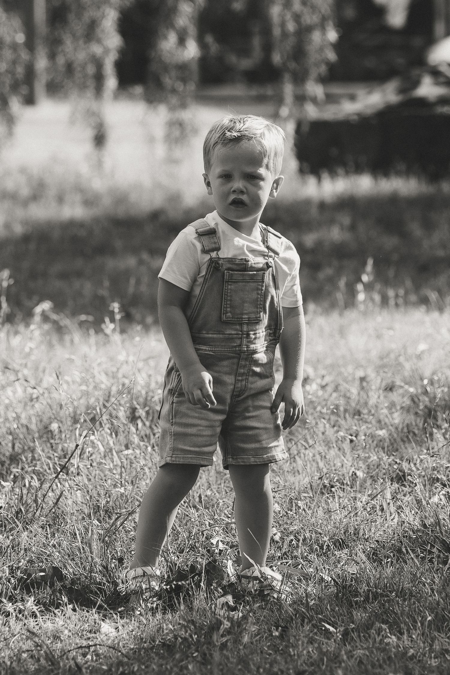 Vintage black and white portrait of small child in overalls standing in grassy field.