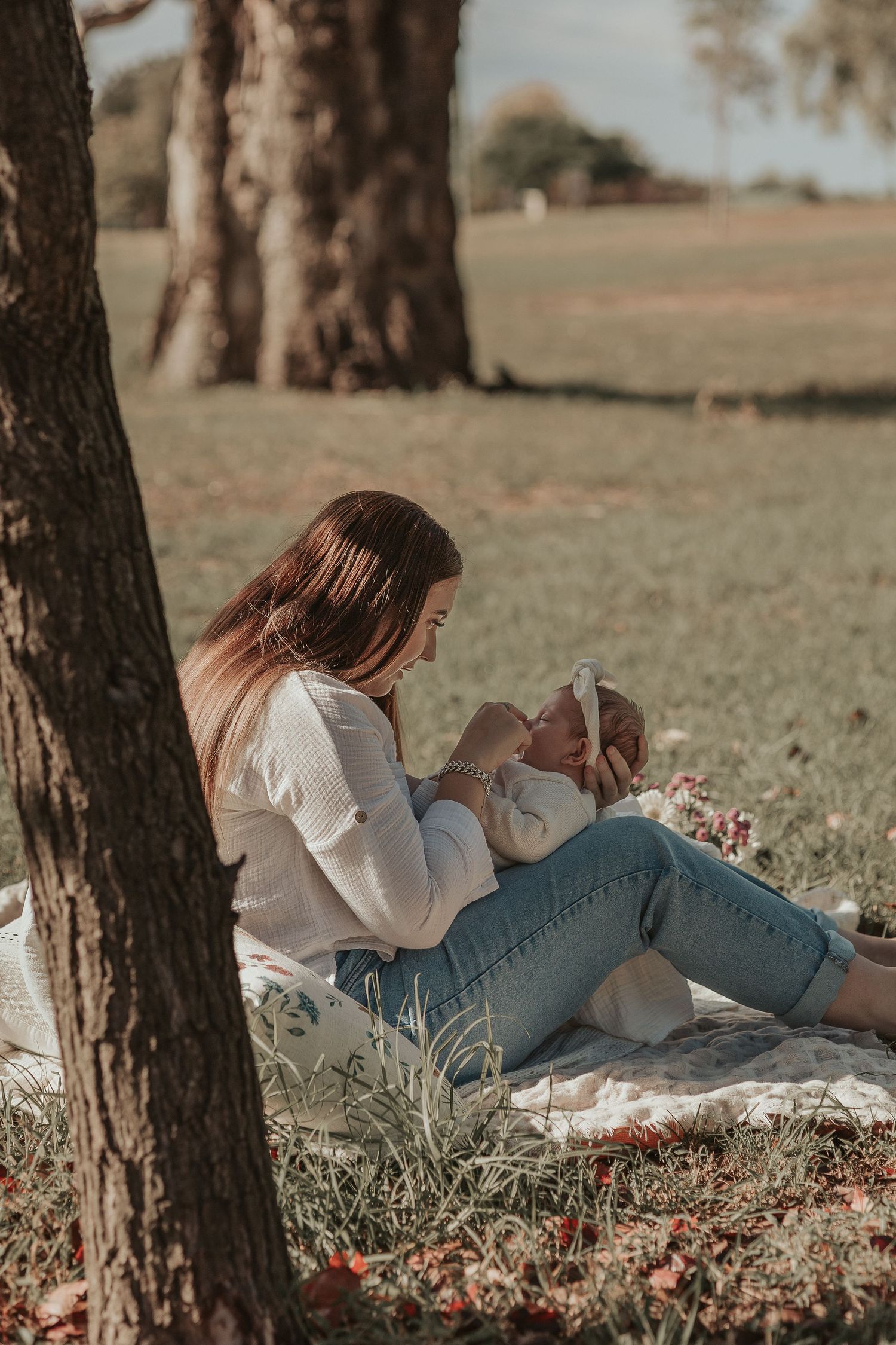 Mother relaxing on blanket holding baby under trees in countryside setting.