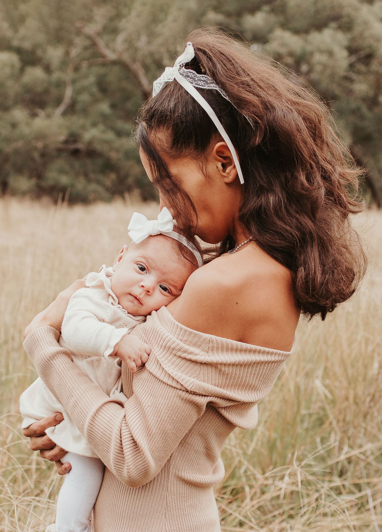 A young mother with dark hair holds her newborn baby in a sunlit field, both dressed in soft neutral tones.