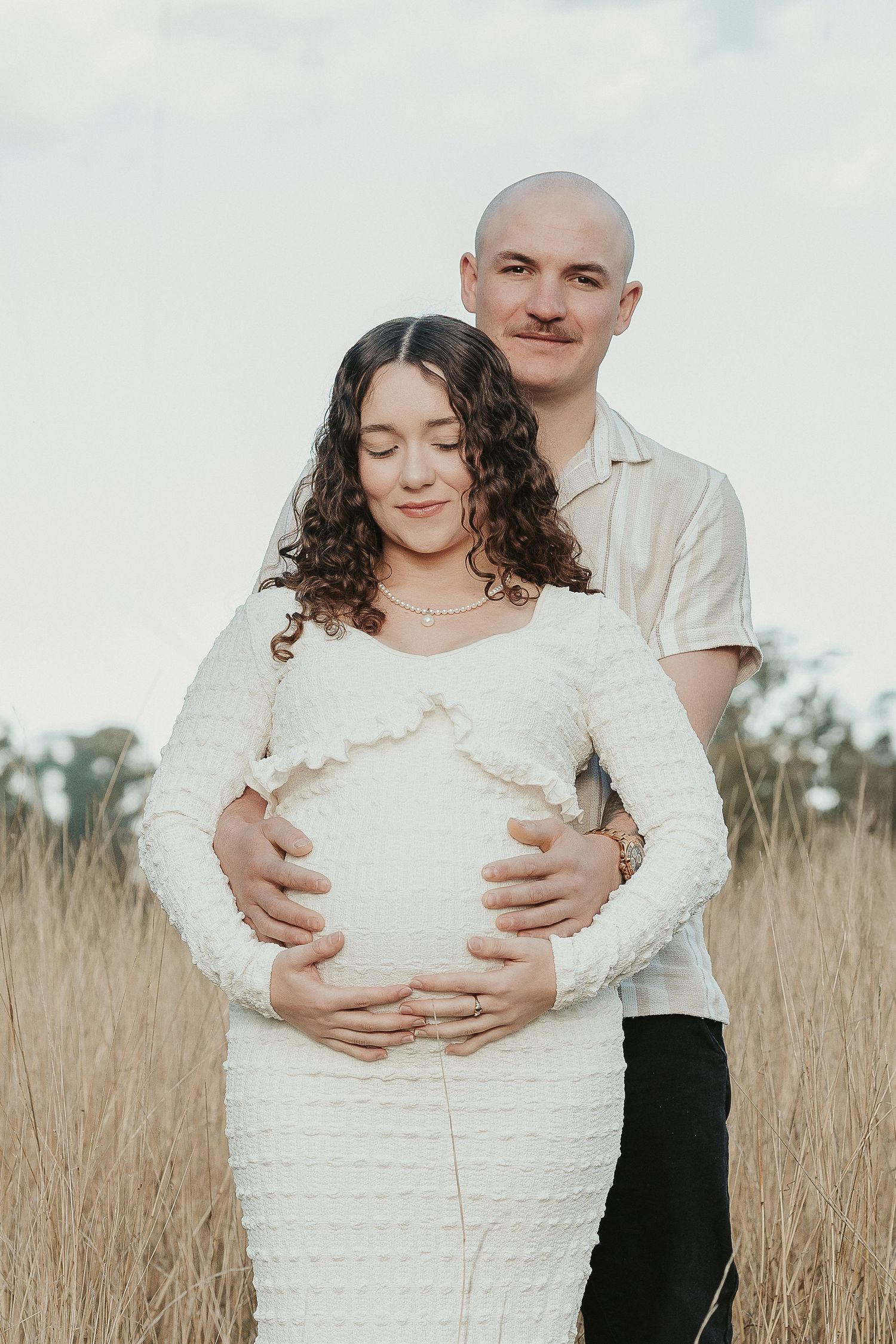 Pregnant woman in white dress embraced by partner in golden field during outdoor maternity photoshoot.