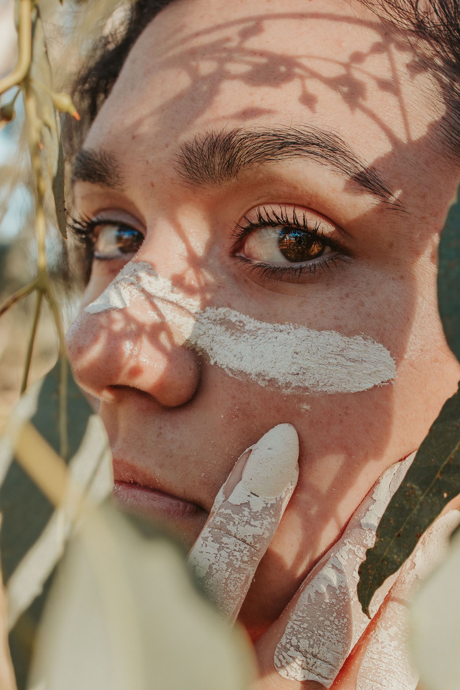 Close-up portrait of a woman with ochre on her face, with natural leaf shadows cast across her skin.