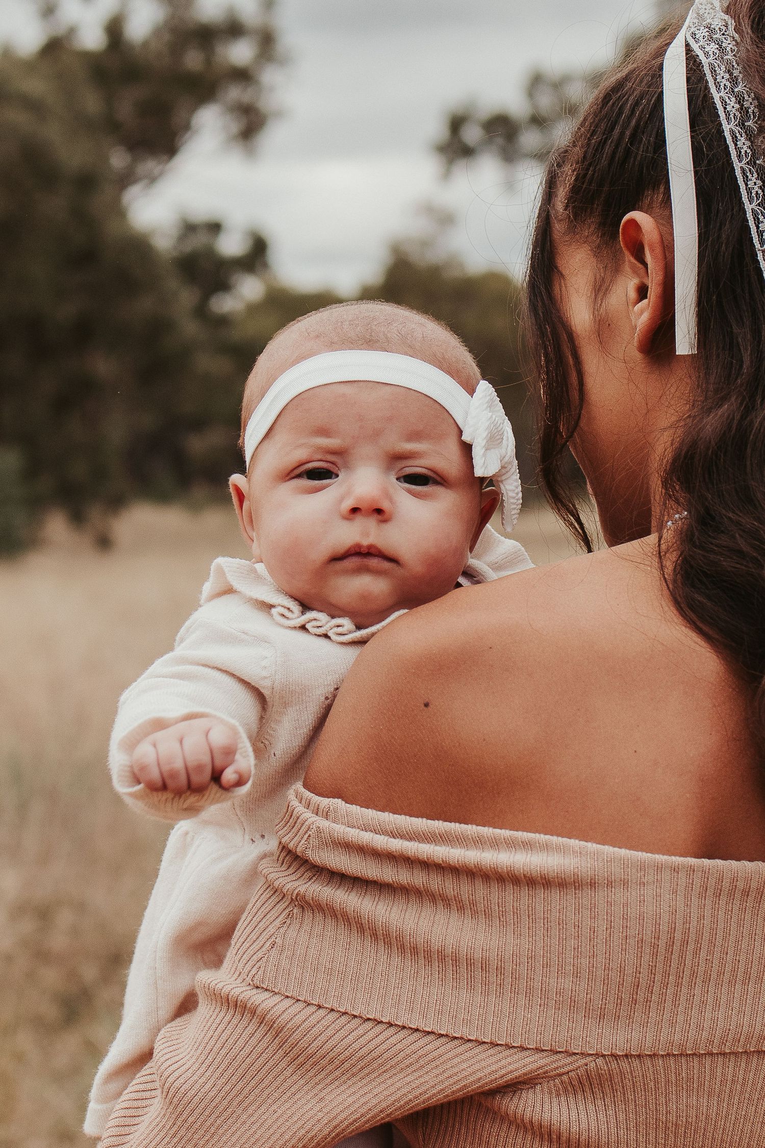 Baby wearing white headband rests on shoulder of person in beige sweater against natural outdoor setting.