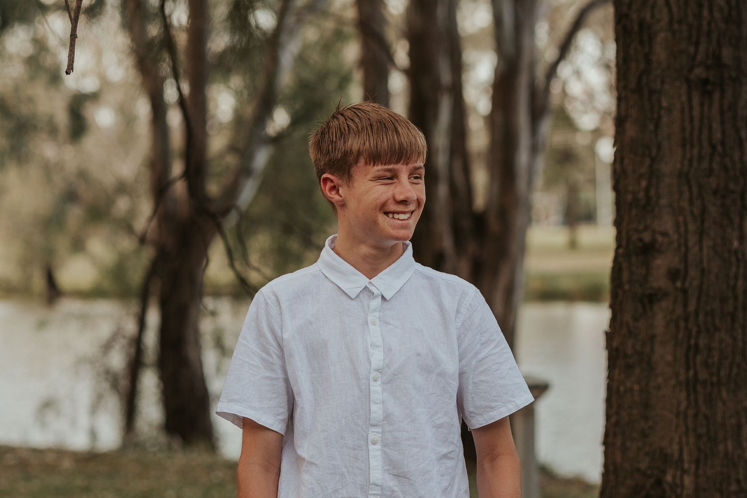 Portrait of young person in white button-up shirt smiling brightly in wooded setting.