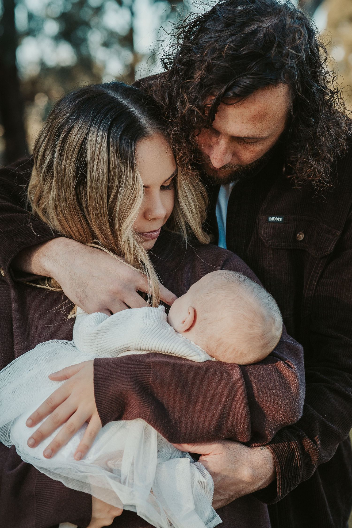 A couple tenderly holds their newborn baby outdoors, surrounded by soft natural light and trees.