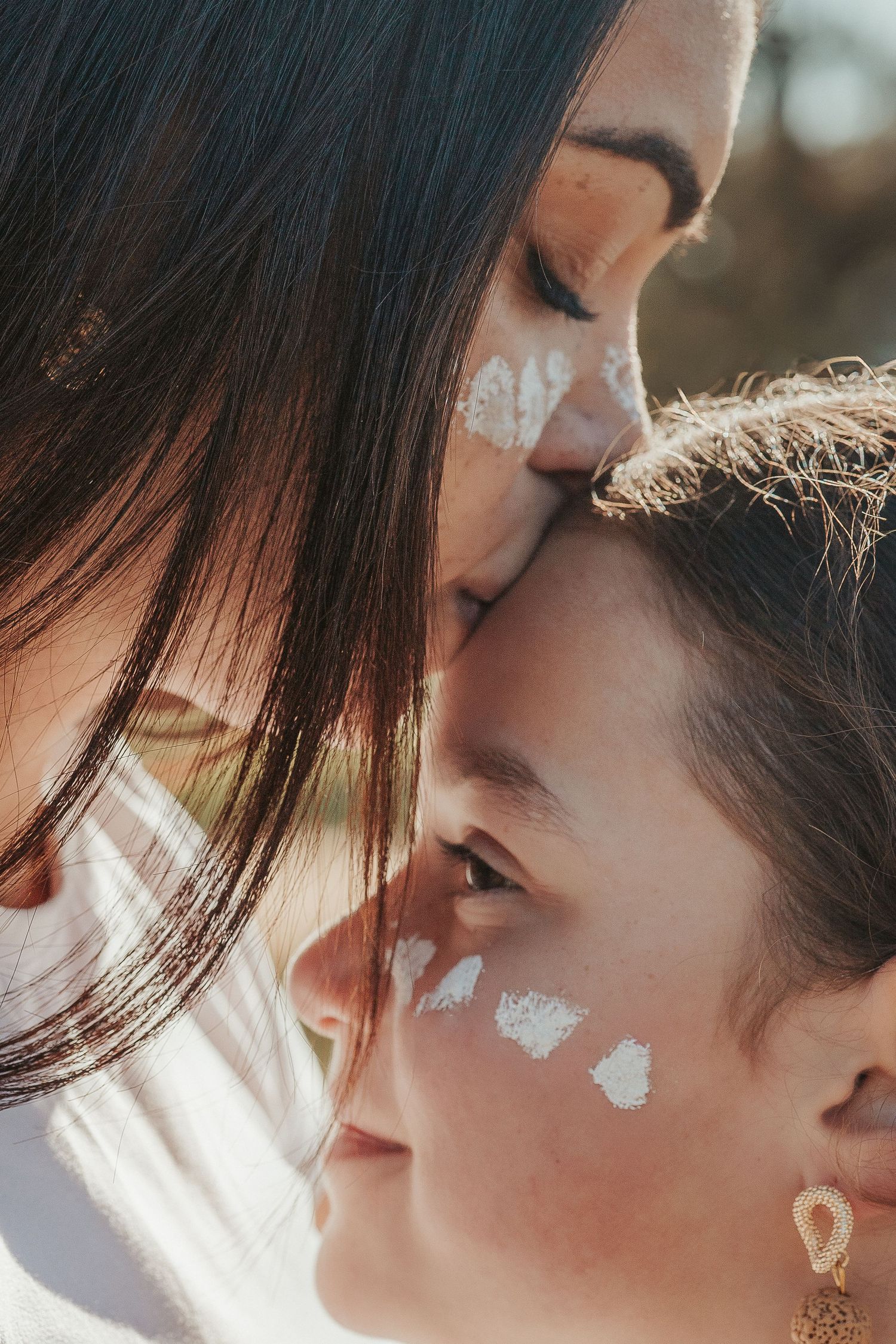 A mother kisses her child's forehead, both with white ochre paint, in a warm sunlit outdoor portrait.