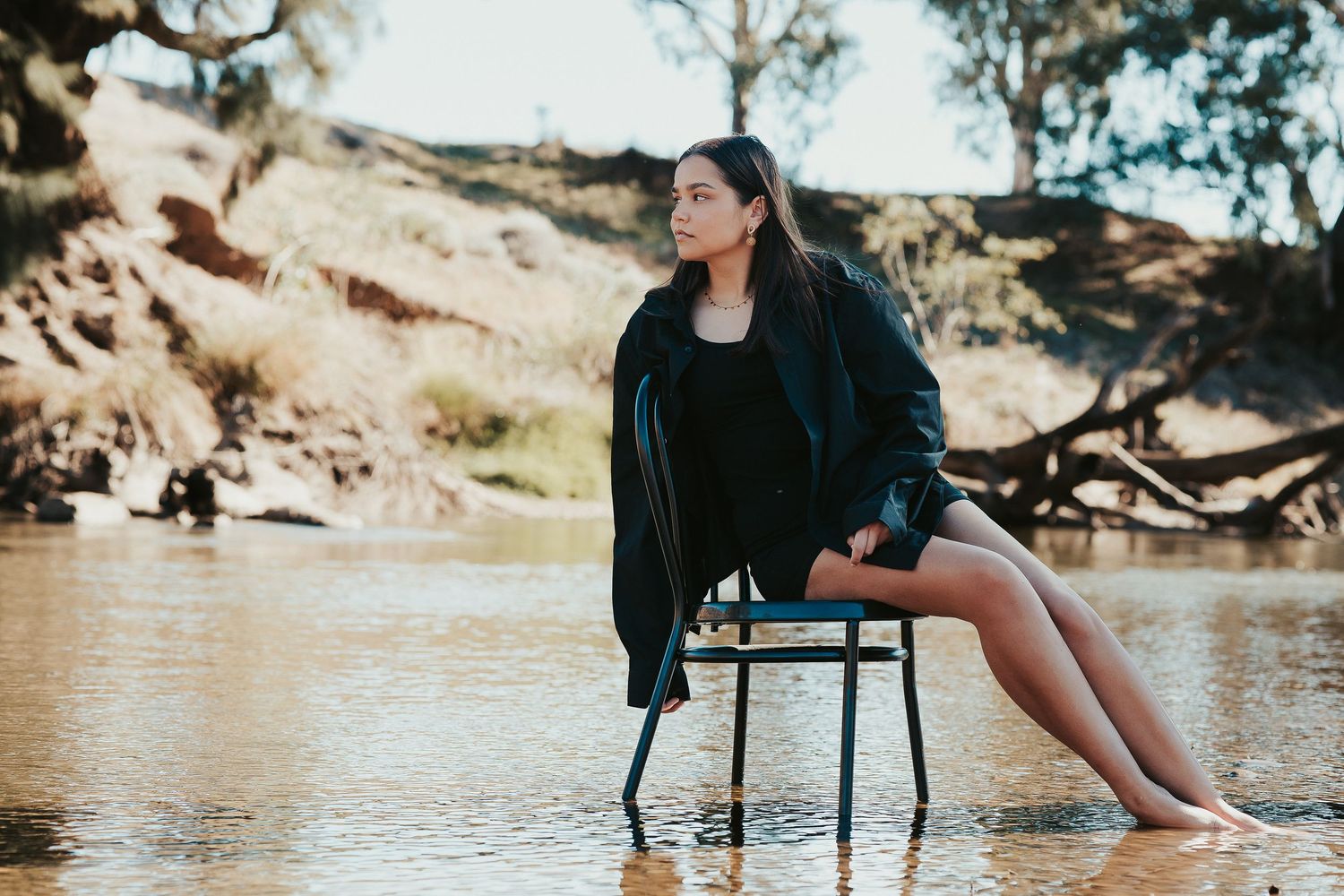 Young woman in black jacket sits on chair in shallow river with sandy banks and trees in background.