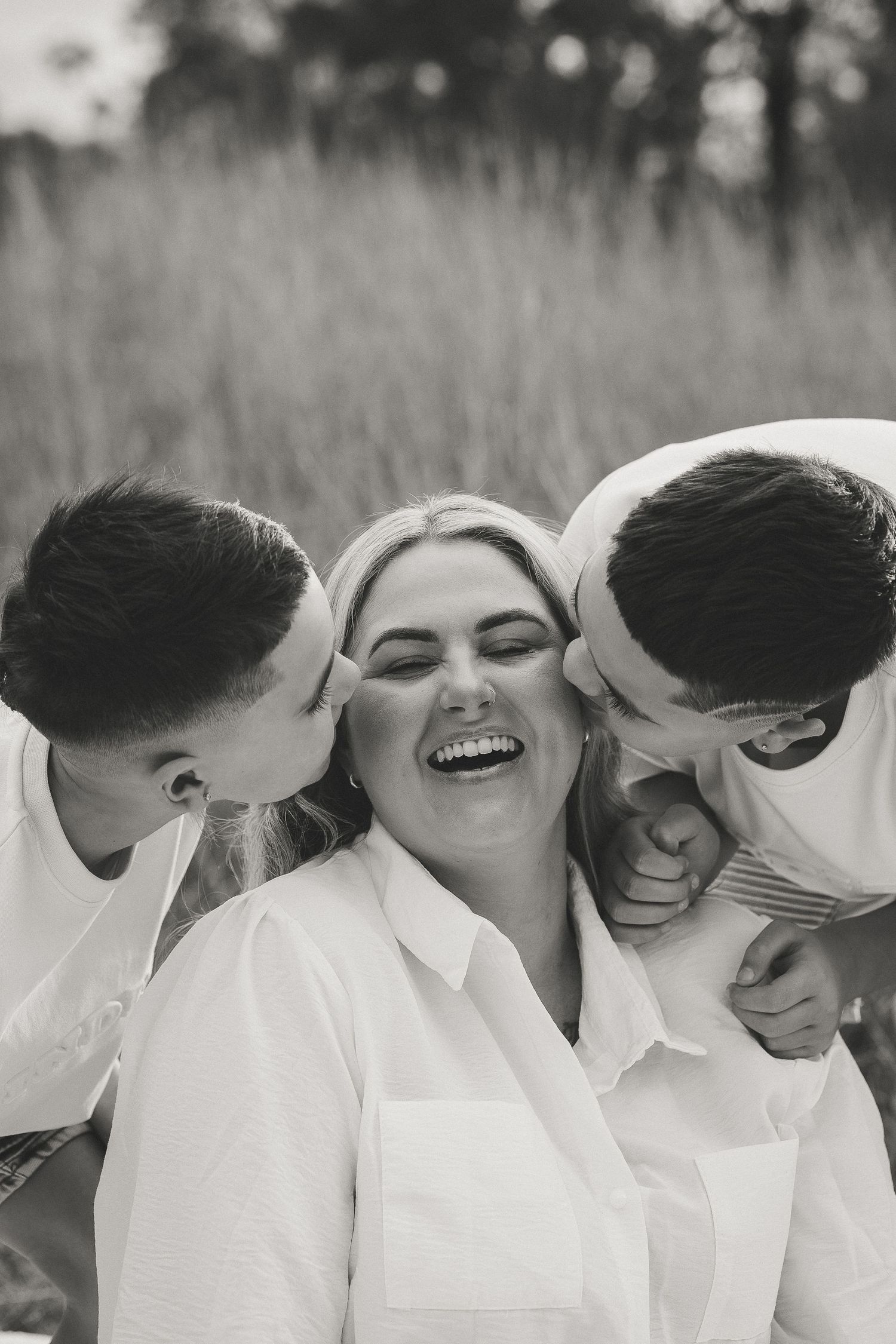Black and white photo of a laughing woman being kissed on both cheeks by two young boys in a field.