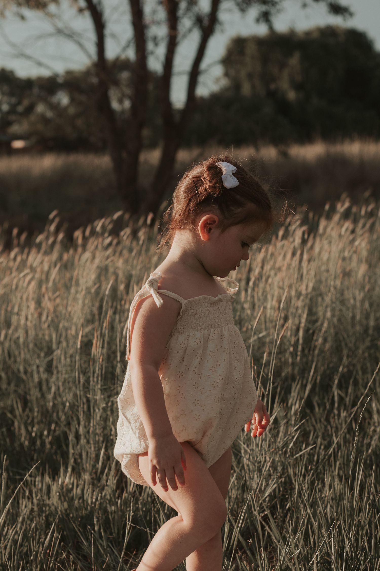 Young child in white dress exploring through tall grass at sunset.