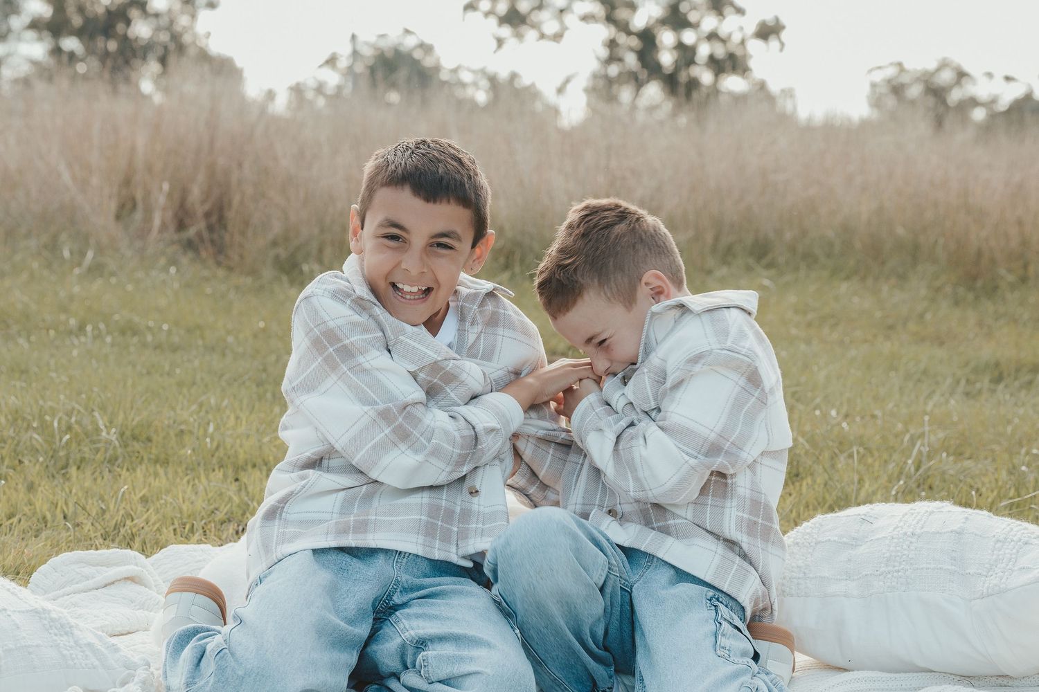 Two young boys in matching plaid shirts sitting on a blanket in a grassy field, smiling and laughing together.