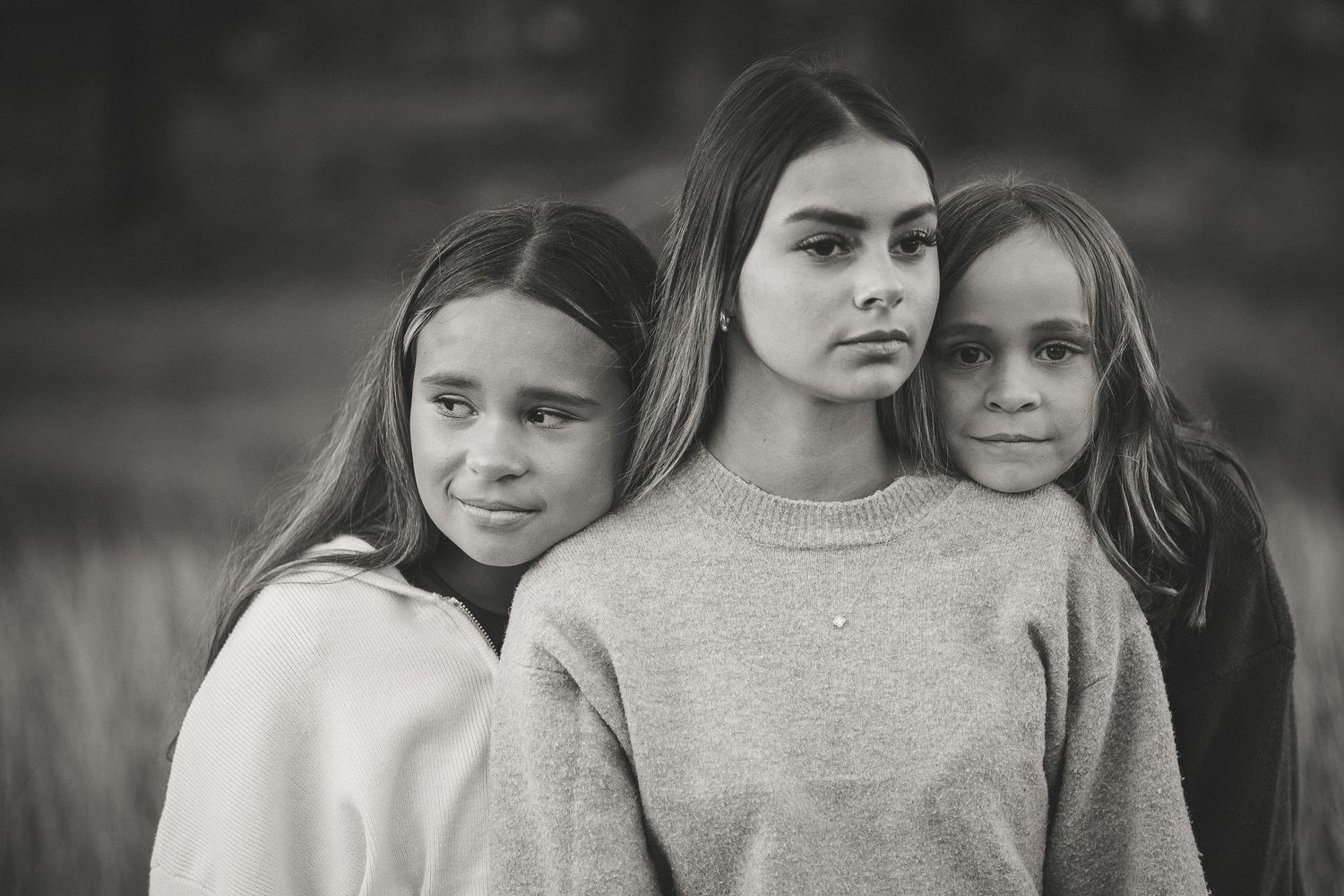 Black and white portrait of three young girls with long hair posing closely together outdoors.