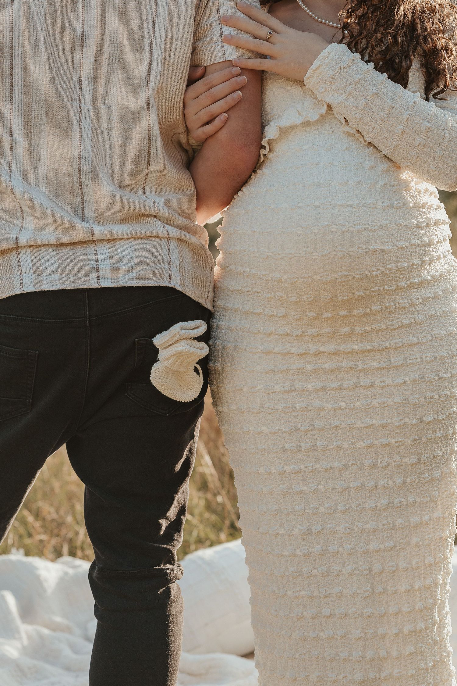 Couple in neutral tones during maternity shoot, tiny baby booties tucked in pocket, pregnant belly visible.