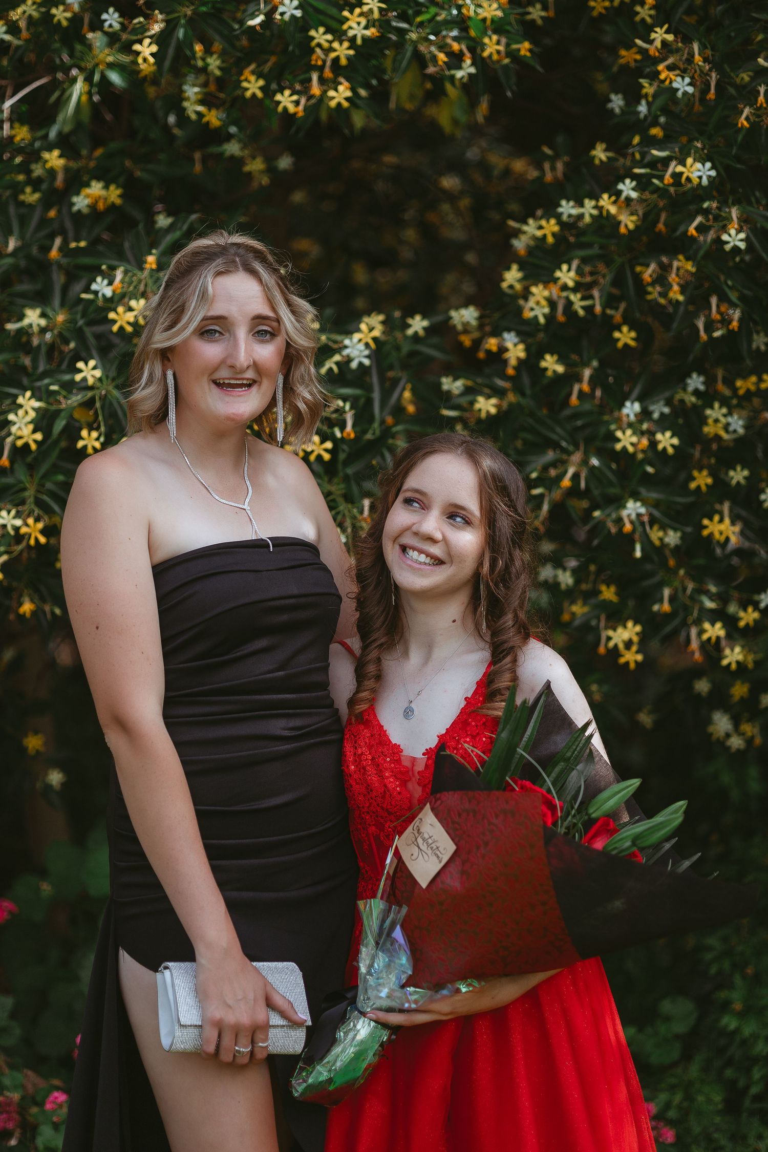 Two people in formal attire, one in black and one in red, pose with flowers against yellow blossoms.