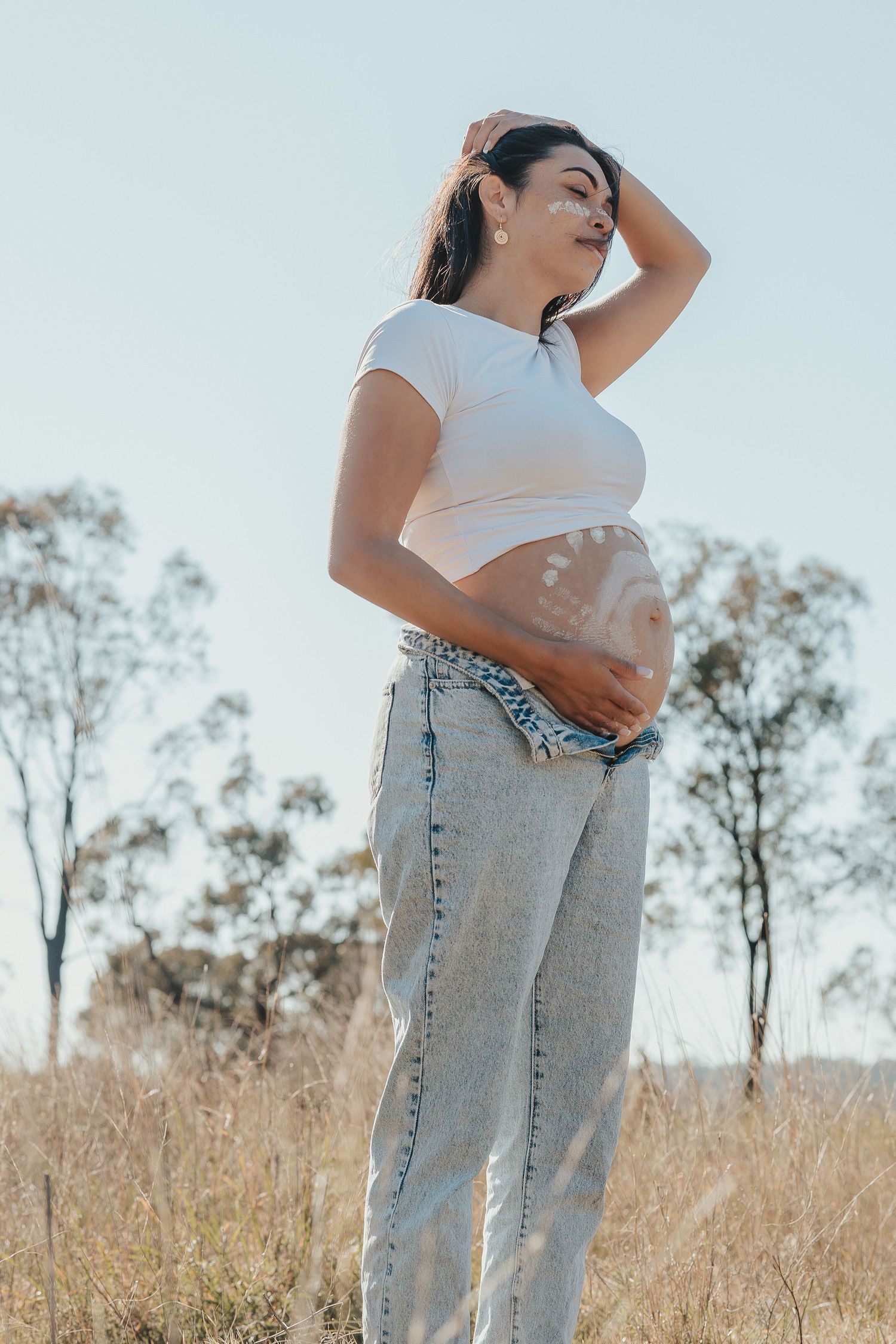 Pregnant woman in white crop top and jeans cradling her baby bump outdoors in a sunny field.