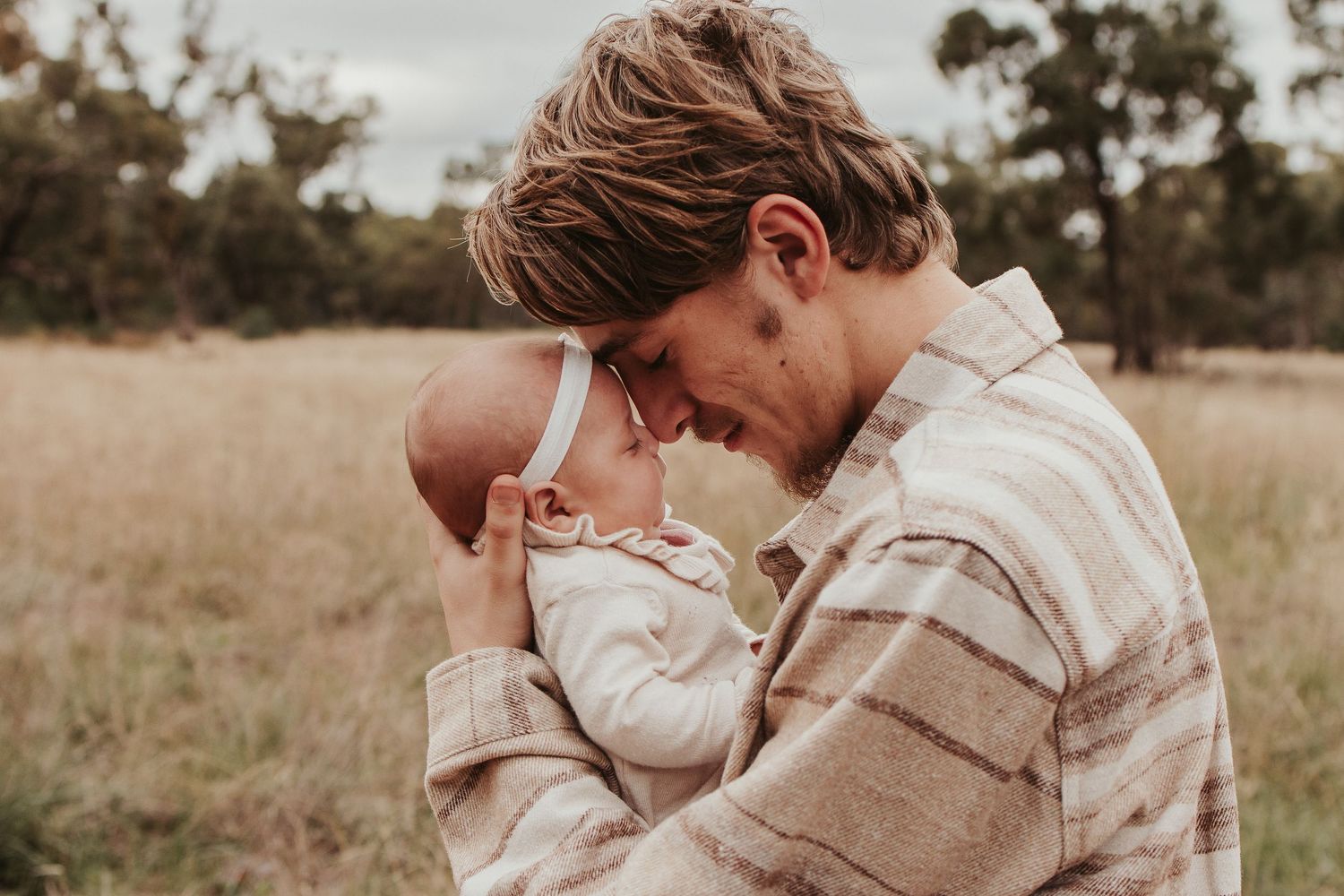 Father tenderly holding newborn baby in a golden field, sharing an intimate forehead-to-forehead moment outdoors.