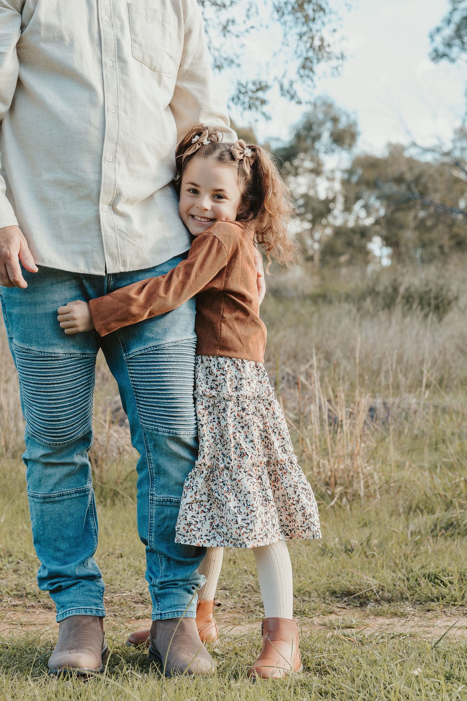 Young girl in floral dress hugs adult's legs outdoors in a grassy field, smiling warmly at camera.