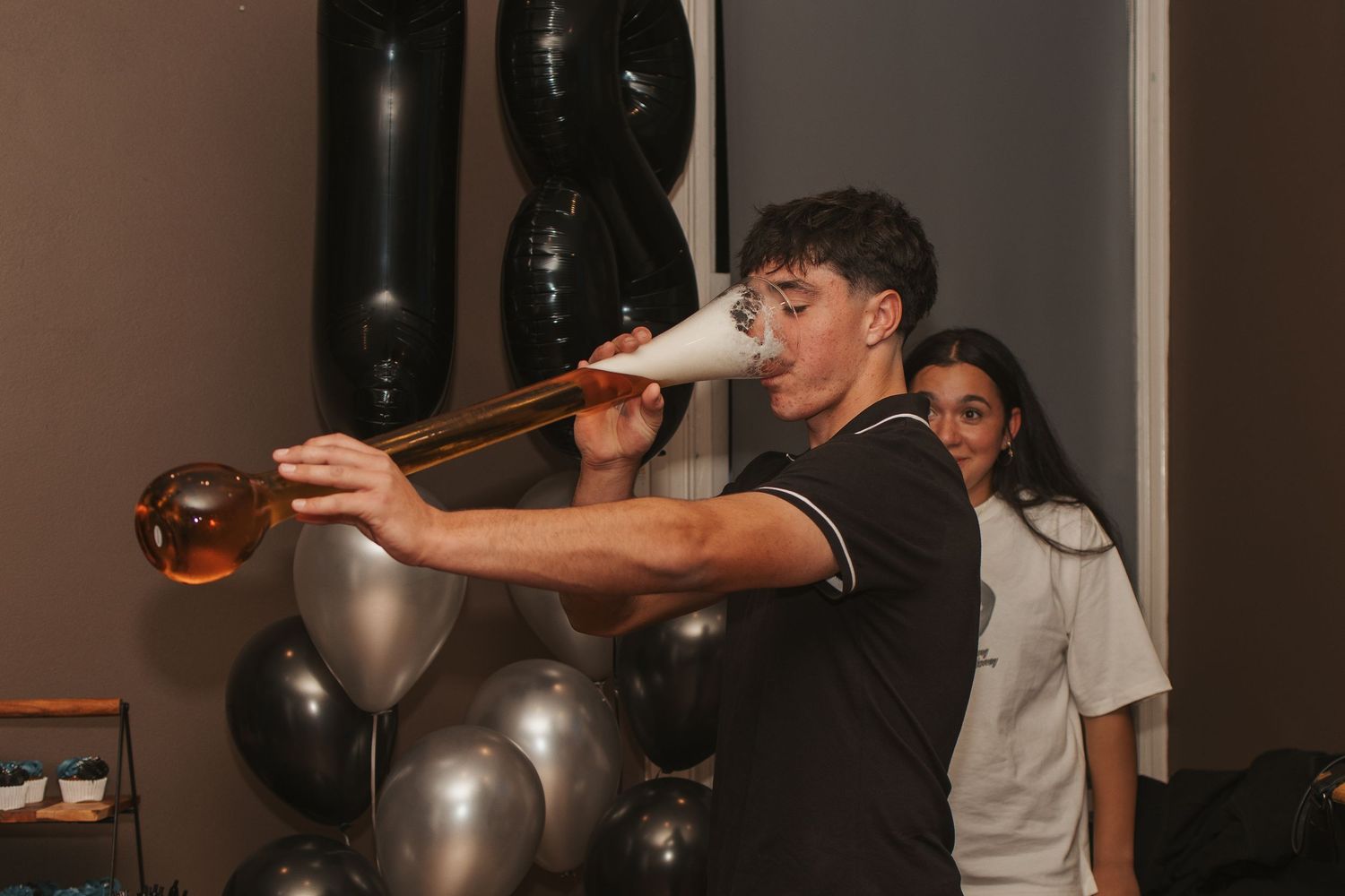 Person drinking from a large bottle at a party with black and silver balloons.