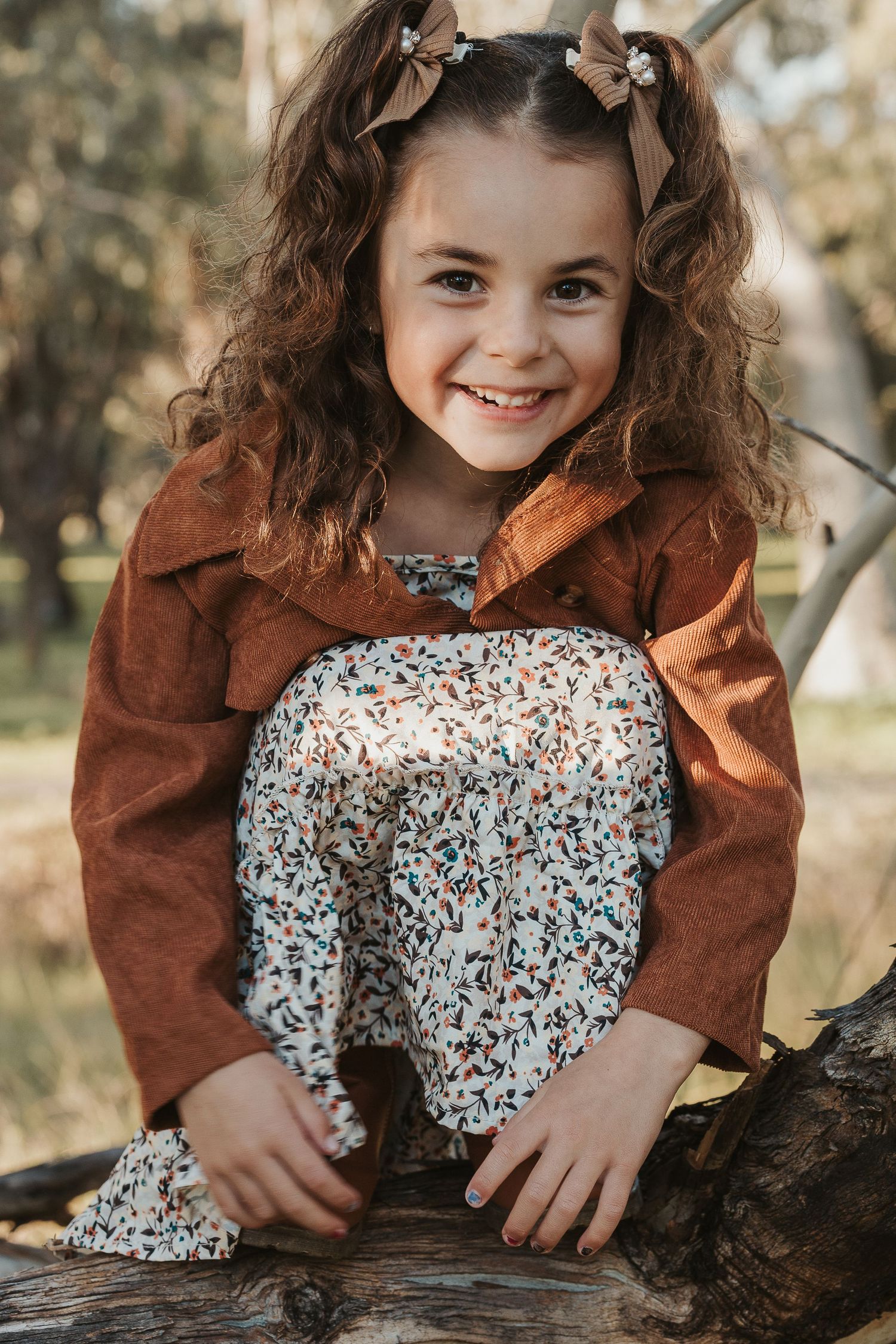 Smiling young girl with curly hair and bows sitting on a log outdoors in a brown jacket and floral pants.