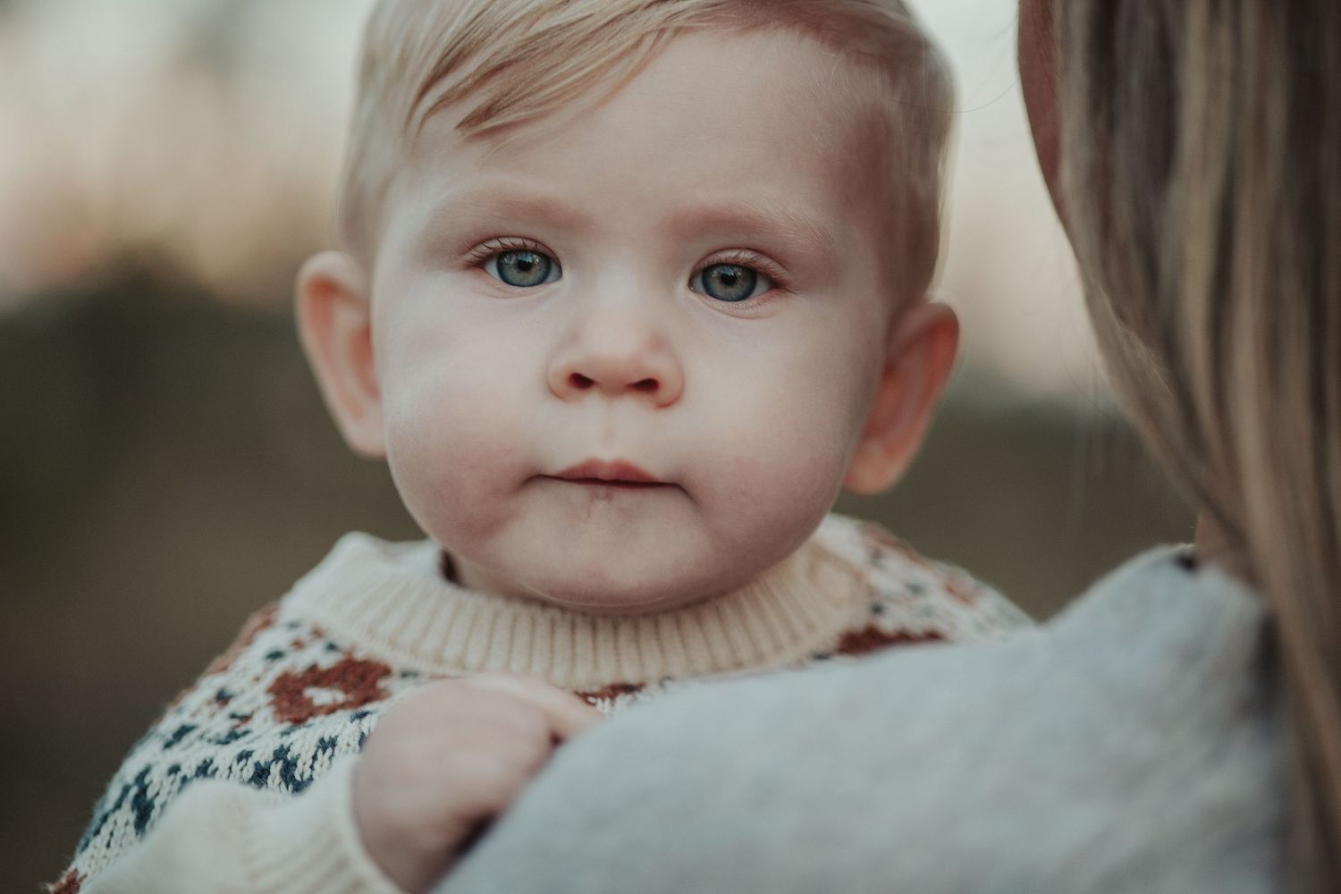 Close-up portrait of a blonde toddler with blue eyes in a knit sweater being held by their mother outdoors.