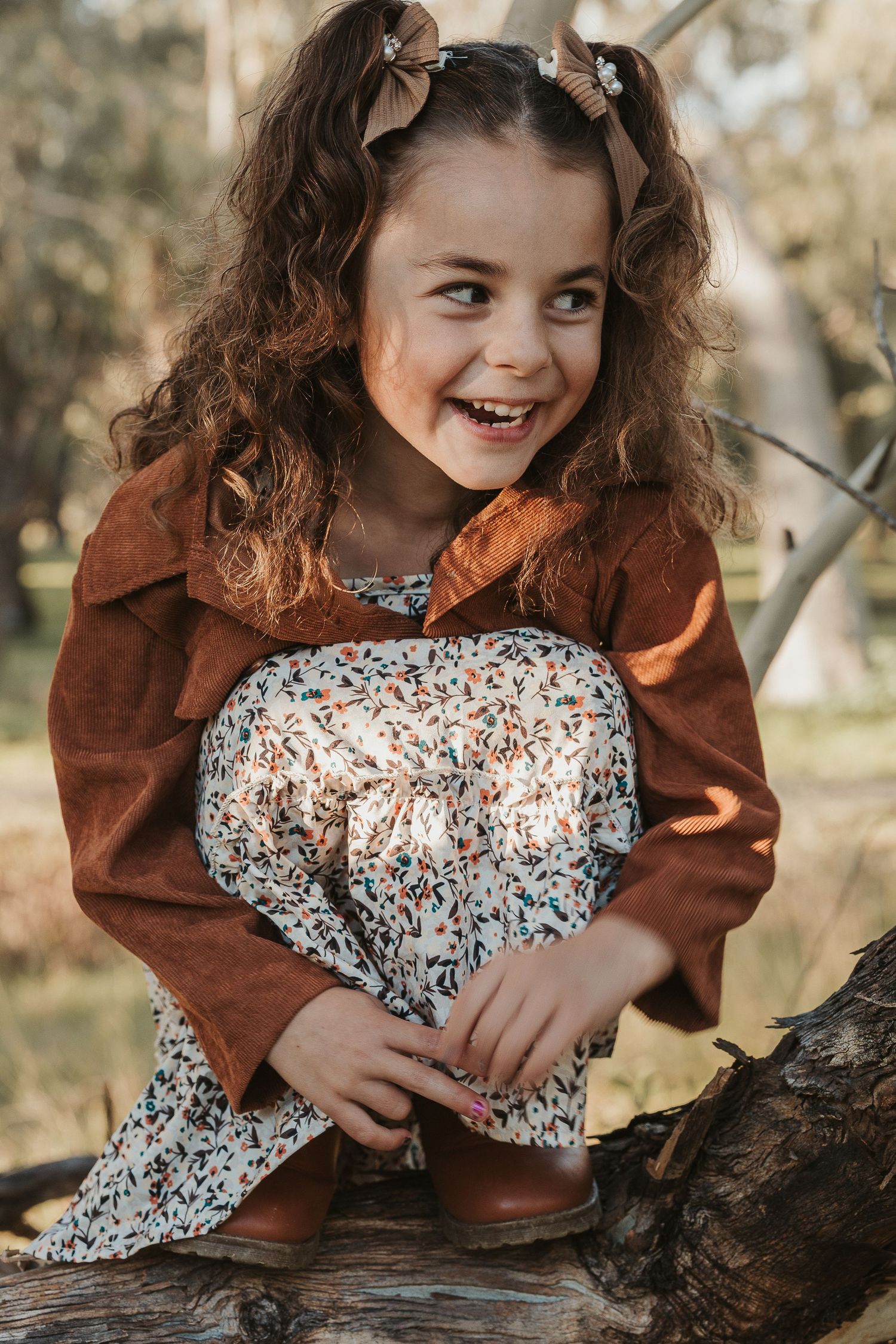 A joyful young girl with curly hair sits on a log in autumn, wearing a floral dress and brown jacket.