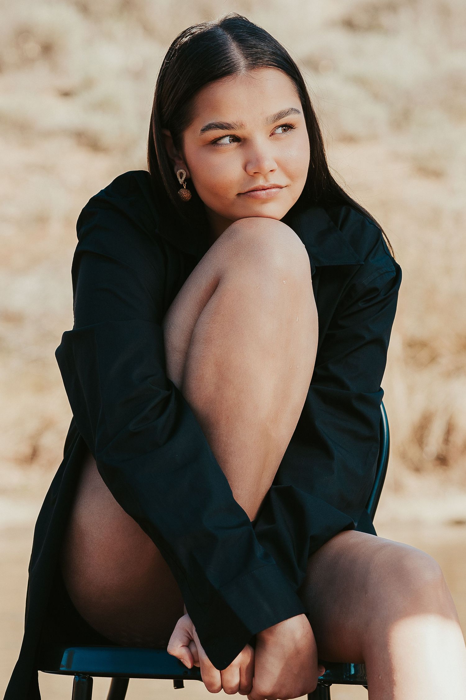 Young woman in black jacket sitting outdoors, hugging her knees, with a sandy background.