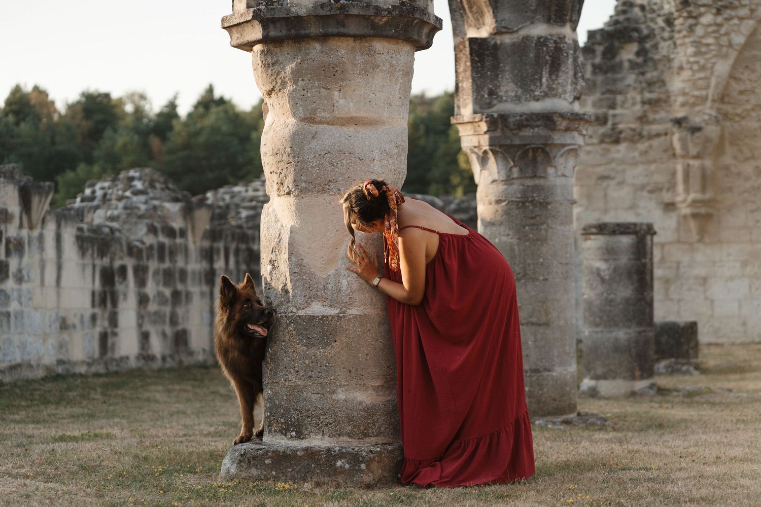 Woman in red dress bending to greet a German Shepherd dog amid ancient stone ruins at dusk.
