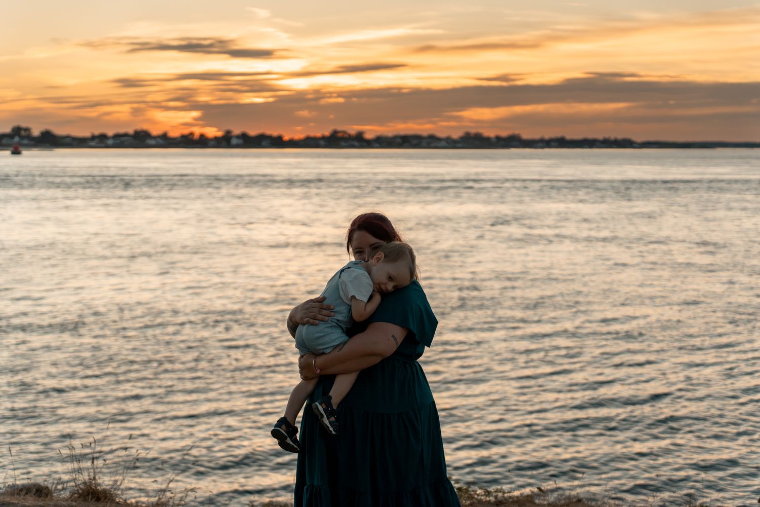 Mother holding baby at golden hour sunset by the water, wearing a teal dress in a tender embrace.