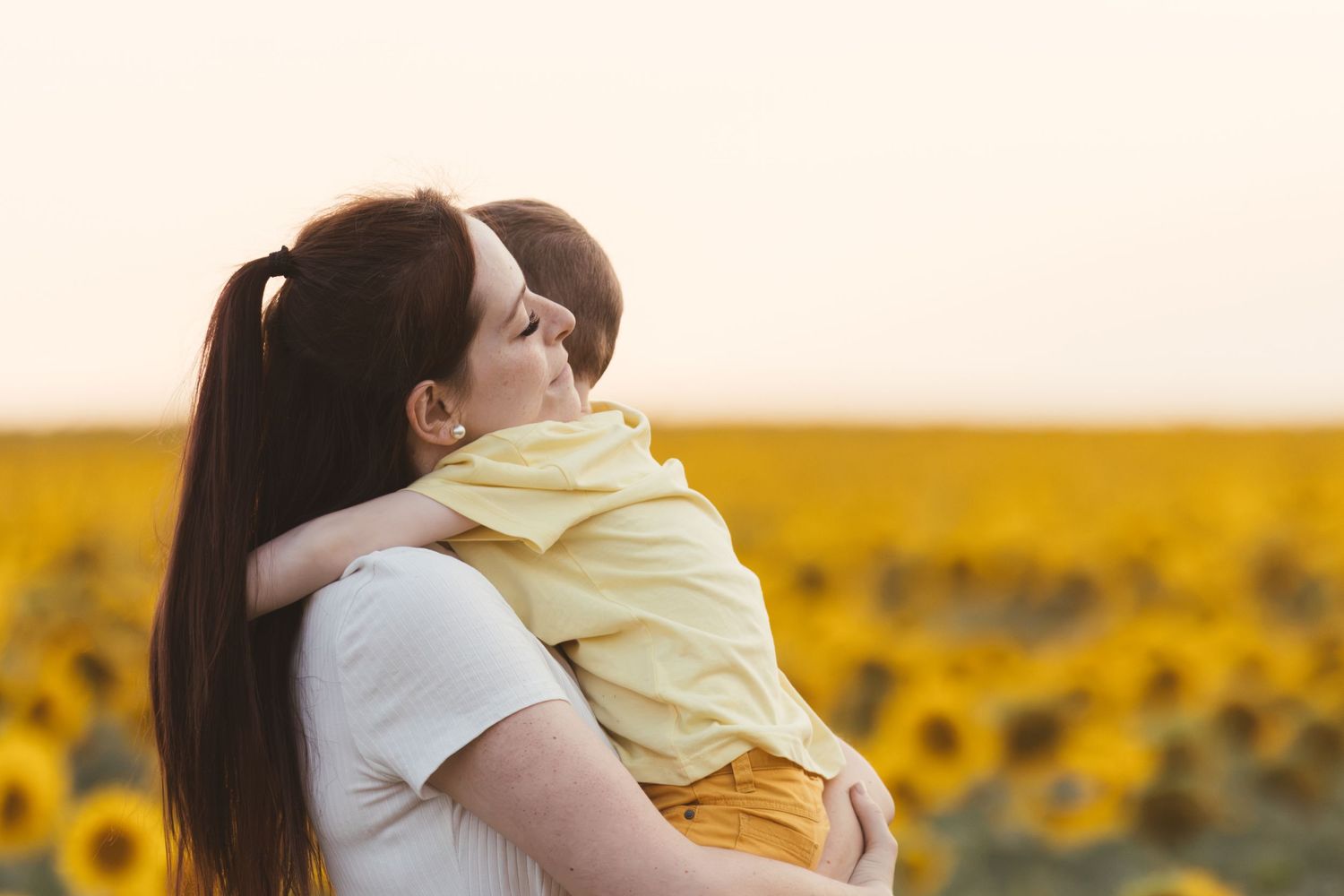 Mother holding young child in sunflower field at sunset, sharing a tender embrace outdoors.