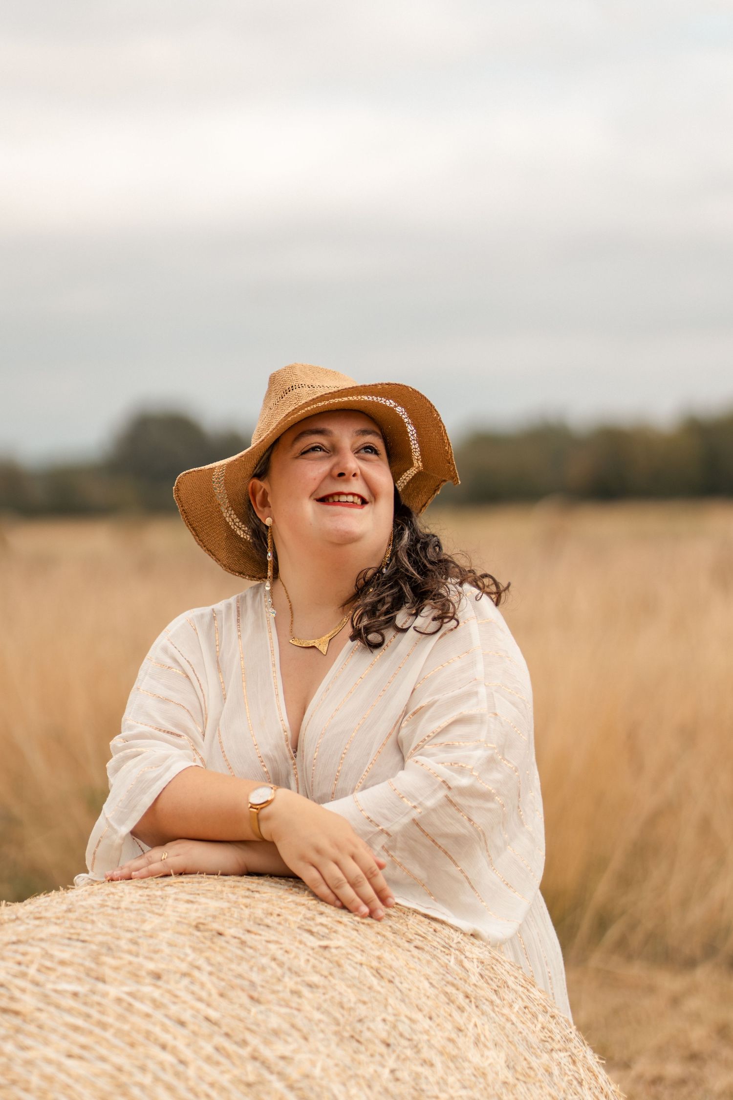 Happy woman in white dress and straw hat leaning on hay bale in field.