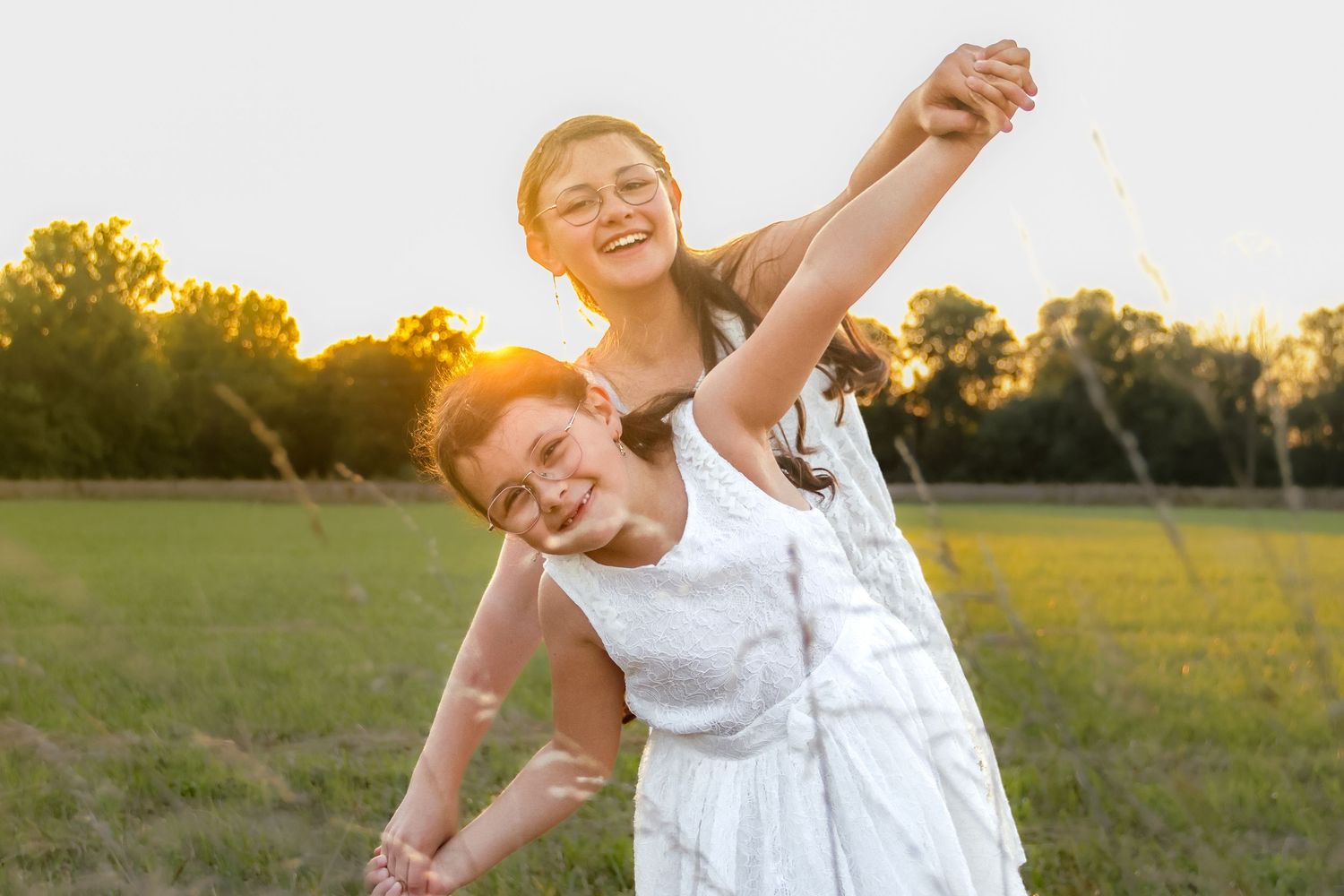 Two smiling girls in white dresses play outdoors in a sunny green field at golden hour.