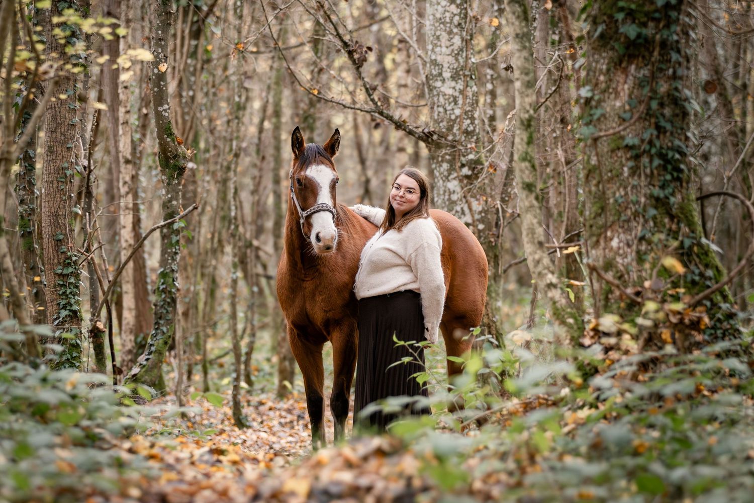 A smiling woman in a white sweater poses with a chestnut horse in a colorful autumn forest.