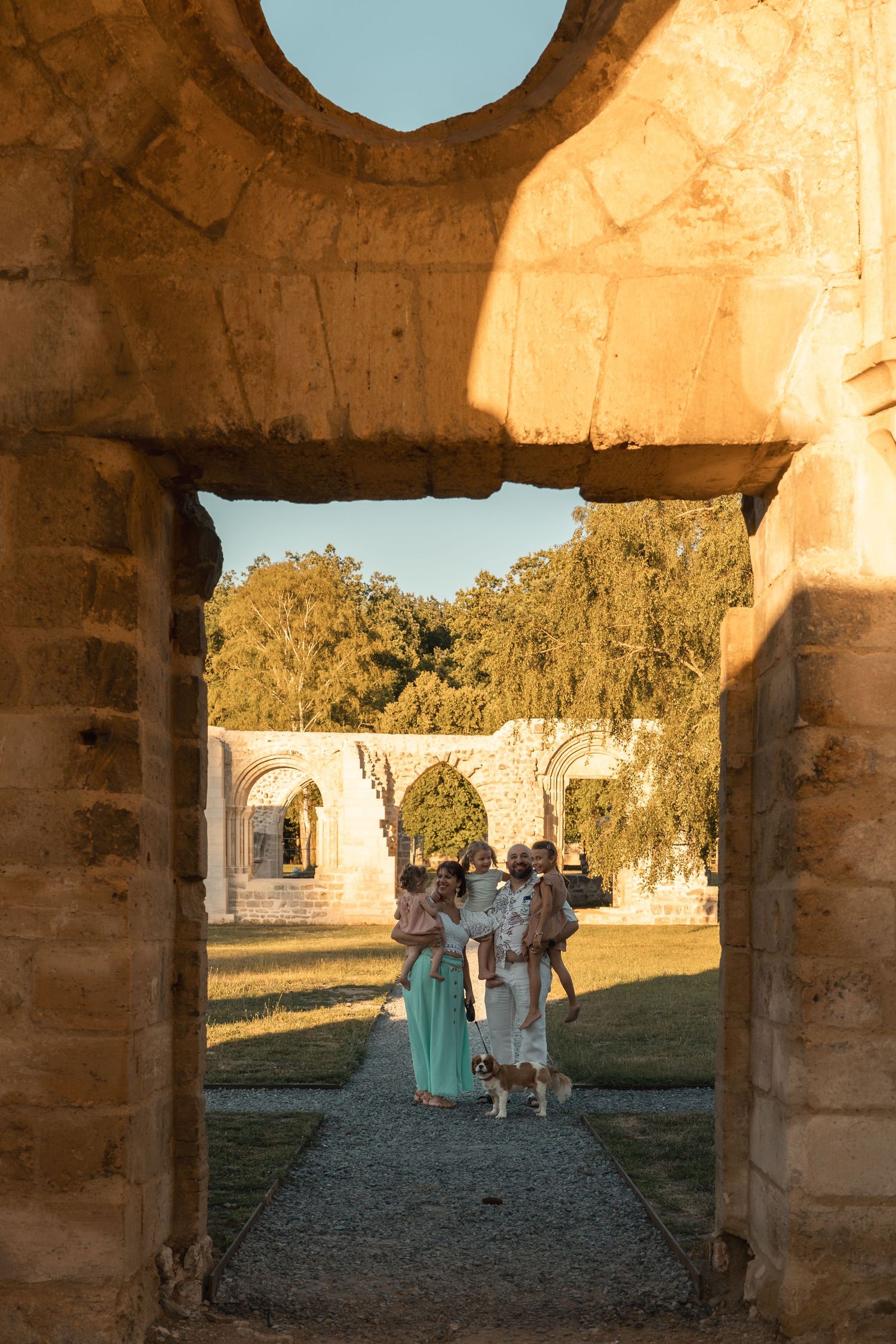 Family posing in ancient stone archway ruins at golden hour, with lush green trees and historic arches in background.