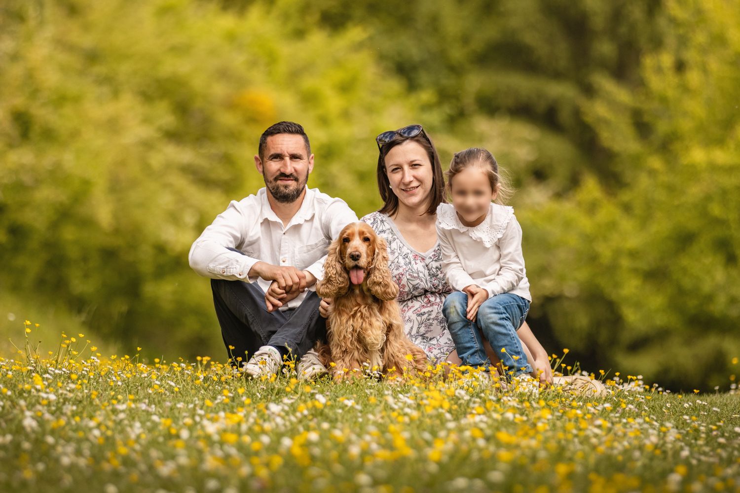 Happy family with a golden Cocker Spaniel sitting in a sunny wildflower meadow surrounded by lush green trees.