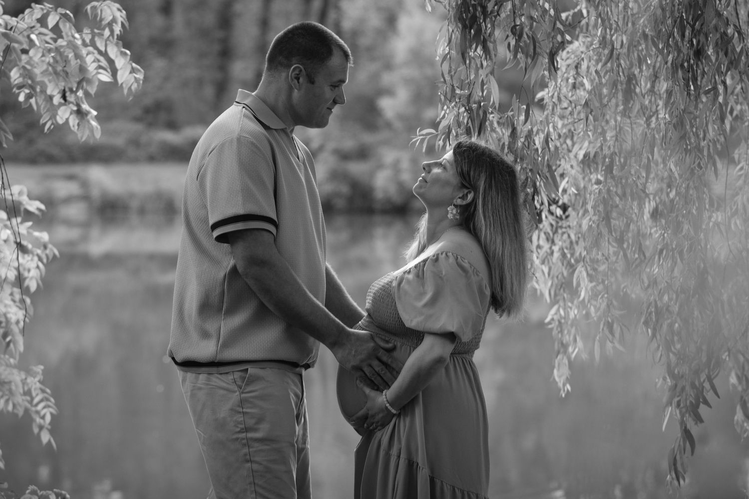 Black and white photo of a couple holding hands and gazing at each other under a willow tree near a lake.